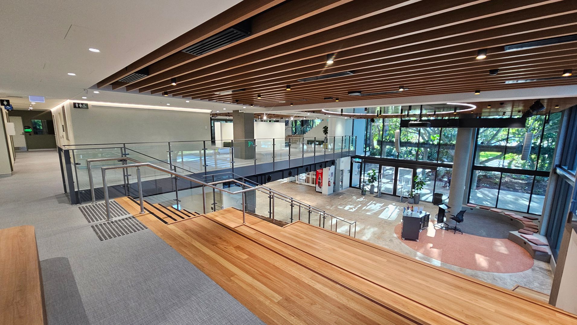 A modern interior space with a wooden staircase leading down to a lower level. The upper level has a grey carpeted floor and glass railings. The ceiling features wooden slats with recessed lighting. The lower level has large glass windows allowing natural light to illuminate the area, with a view of greenery outside. There is a circular seating area with cushions and a small table with chairs. The overall design is open and airy, with a mix of wood and glass elements.