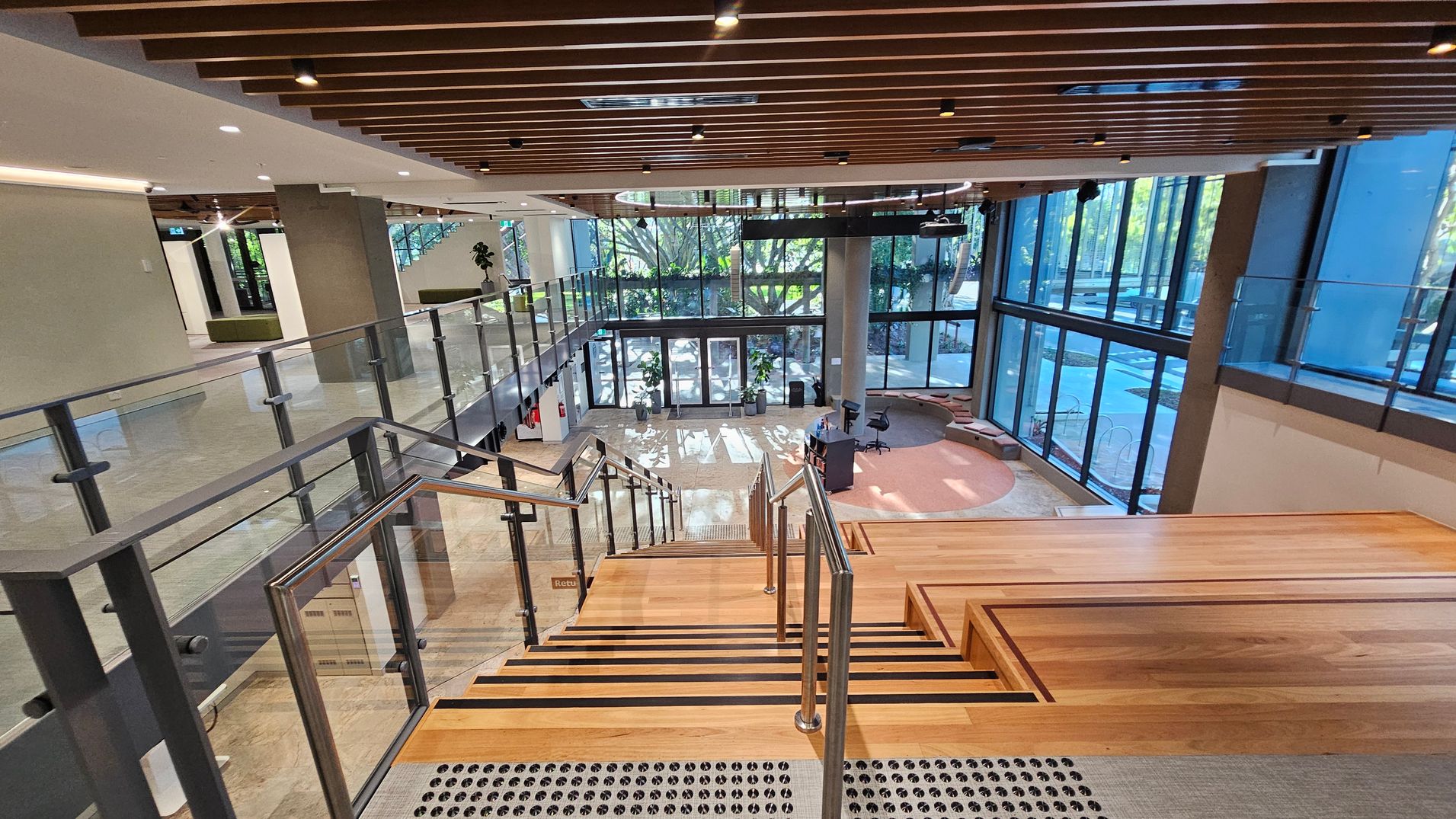 A modern interior space with a wide wooden staircase leading down to a lobby area. The staircase has metal railings and glass panels on the sides. The floor below is tiled, with a circular carpeted area and a small desk with a chair. Large glass windows on the right allow natural light to flood in, revealing greenery outside. The ceiling features wooden slats with recessed lighting. On the left, there is a corridor with more glass panels and a few potted plants.