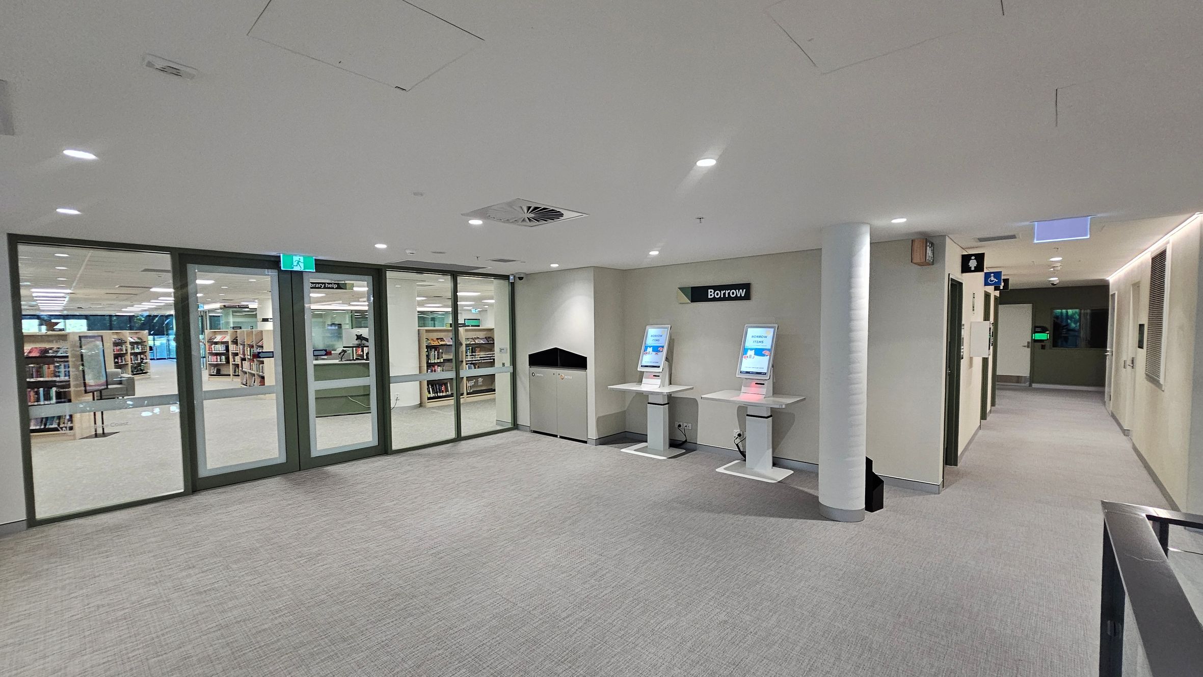 A library entrance area with grey carpeted flooring. On the left, there are large glass doors leading to the main library space, where bookshelves are visible. In the centre, two self-service kiosks with screens are labelled "Borrow". On the right, a corridor leads to restrooms, indicated by signage. The ceiling is white with recessed lighting and ventilation panels.