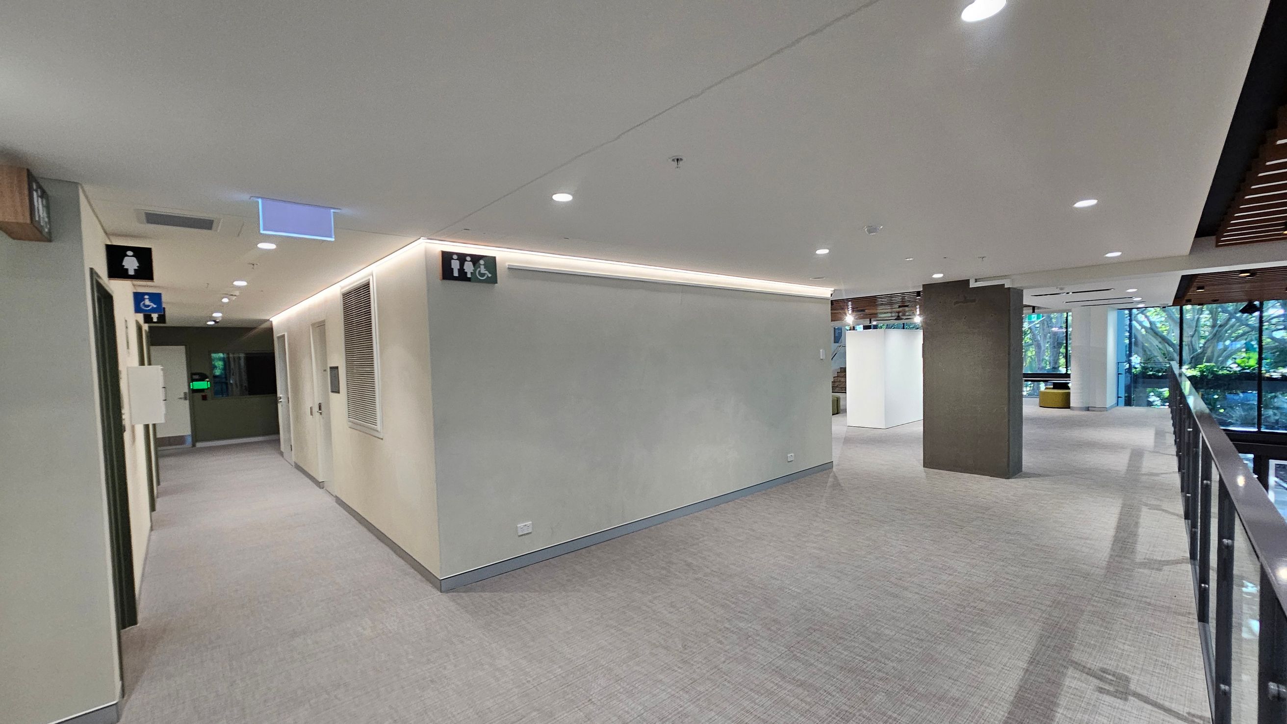 A modern indoor corridor with grey carpeted flooring and a white ceiling with recessed lighting. On the left, there are doors with signs indicating male, female, and accessible toilets. The corridor leads to a darker area with a green-lit exit sign. On the right, a glass railing overlooks a lower level, and large windows reveal greenery outside. A grey column stands near the centre, and the ceiling features wooden slats near the windows.