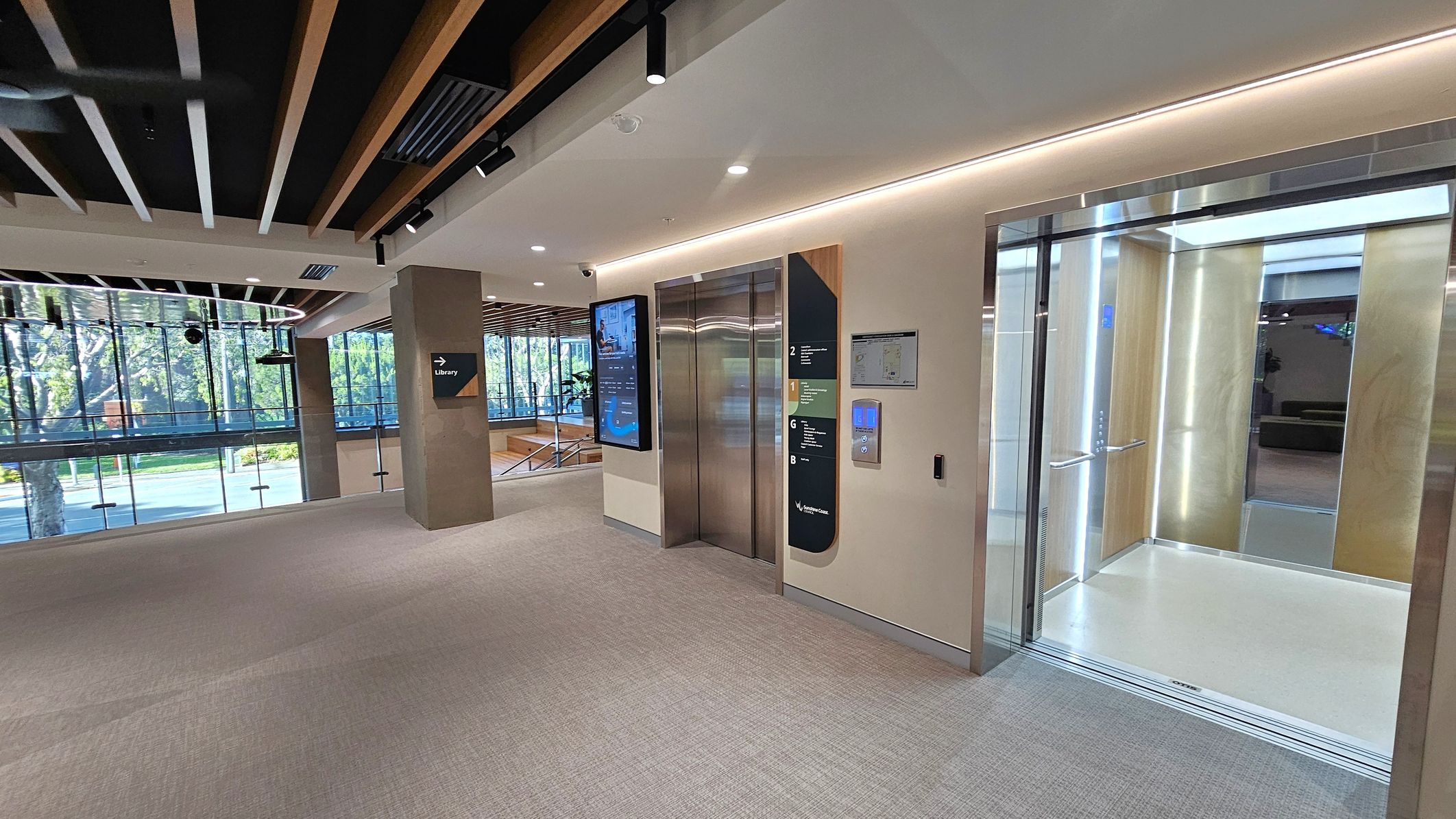 A modern interior with two lifts on the right, one open and one closed. The open lift has a bright interior with wood panelling and LED lighting. A digital directory and control panel are mounted on the wall between the lifts. On the left, large windows offer a view of trees outside. A sign on a pillar points towards the library. The ceiling features wooden slats and recessed lighting, while the floor is carpeted in a neutral tone.