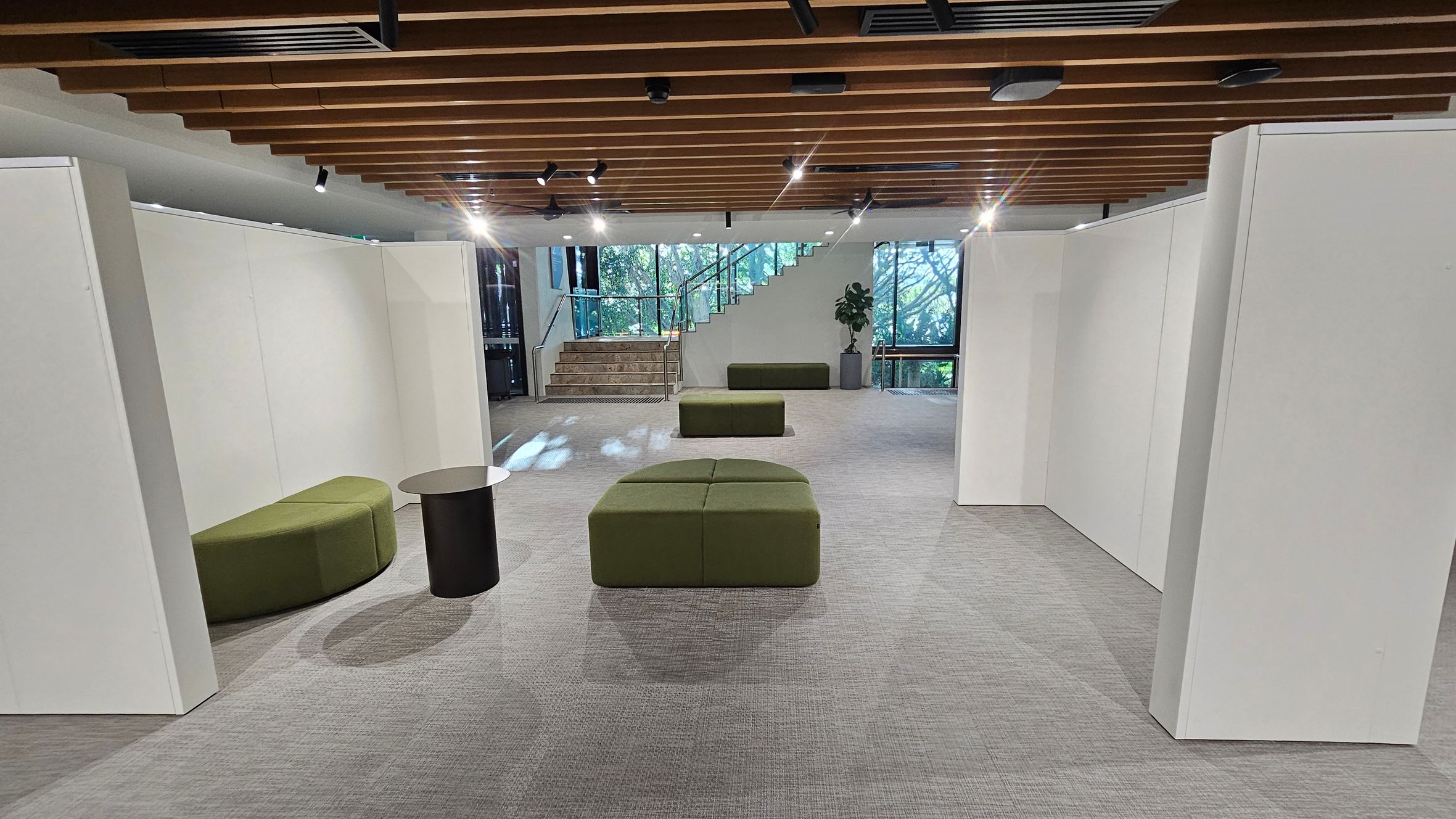 A modern interior space with grey carpeted flooring and a wooden slatted ceiling. In the centre, there are green cushioned benches and a small round black table. On the left, a curved green bench is positioned against a white partition wall. At the back, a staircase with glass railings leads upwards, flanked by large windows showing greenery outside. A potted plant is placed near the stairs. The lighting is provided by spotlights on the ceiling.