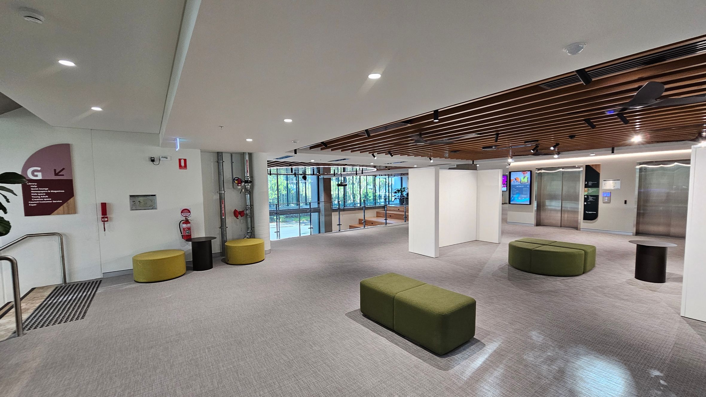 A modern interior space with a grey carpeted floor. On the left, there are yellow circular seats and a small black table near a wall with a sign and fire safety equipment. In the centre, green rectangular seats are placed on the floor. The right side features two elevators with digital screens nearby. The ceiling has wooden slats and recessed lighting. Large windows in the background allow natural light to enter, with a view of greenery outside.