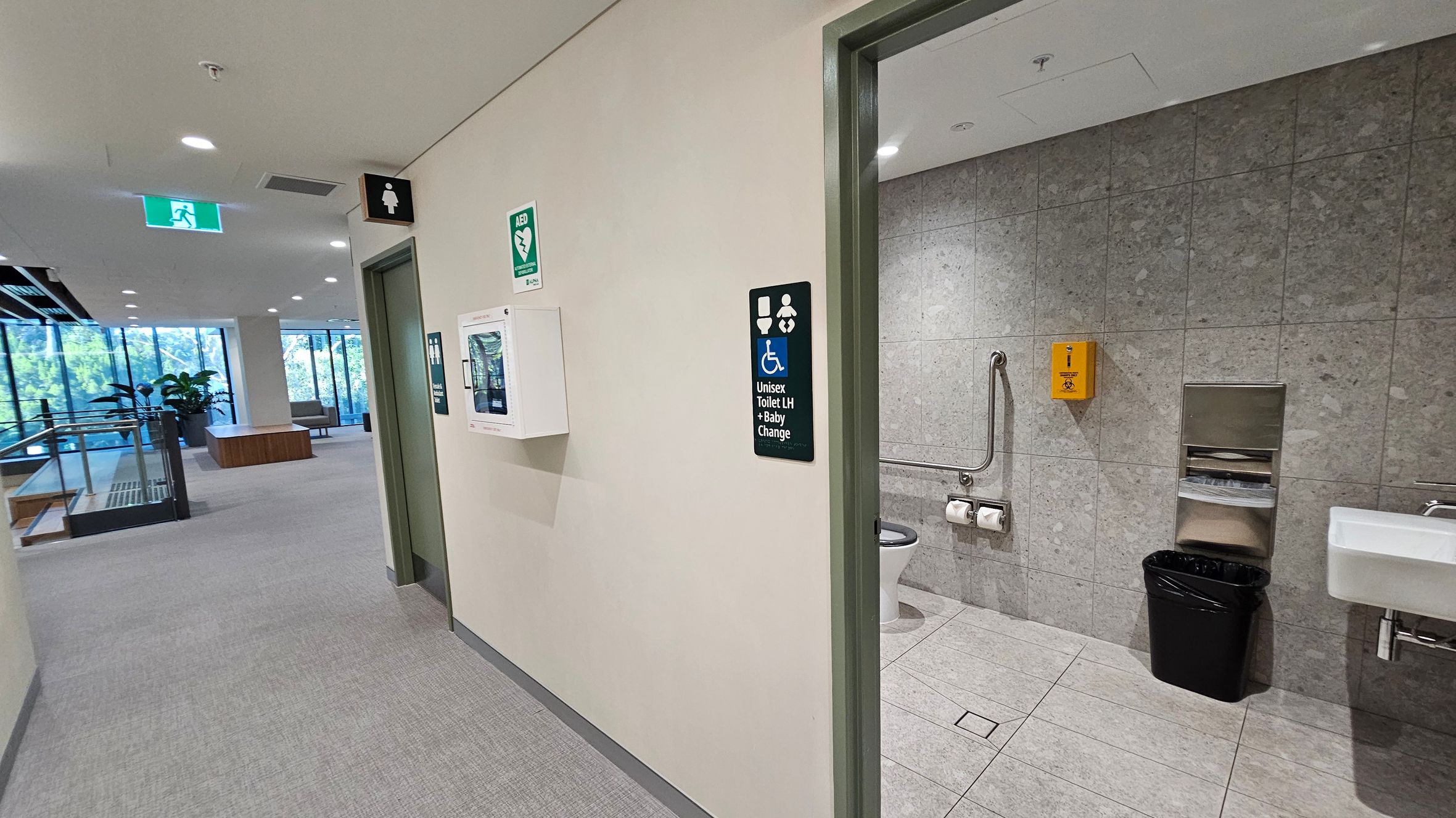 A hallway with a unisex toilet on the right. The toilet has grey tiled walls, a grab bar, toilet paper, a yellow disposal unit, a black bin, and a sink. Signs indicate accessibility and baby changing facilities. An AED unit is mounted on the wall outside. The hallway has a grey carpet, ceiling lights, and an emergency exit sign. Large windows and a staircase with glass railings are visible in the background, along with some indoor plants and seating.