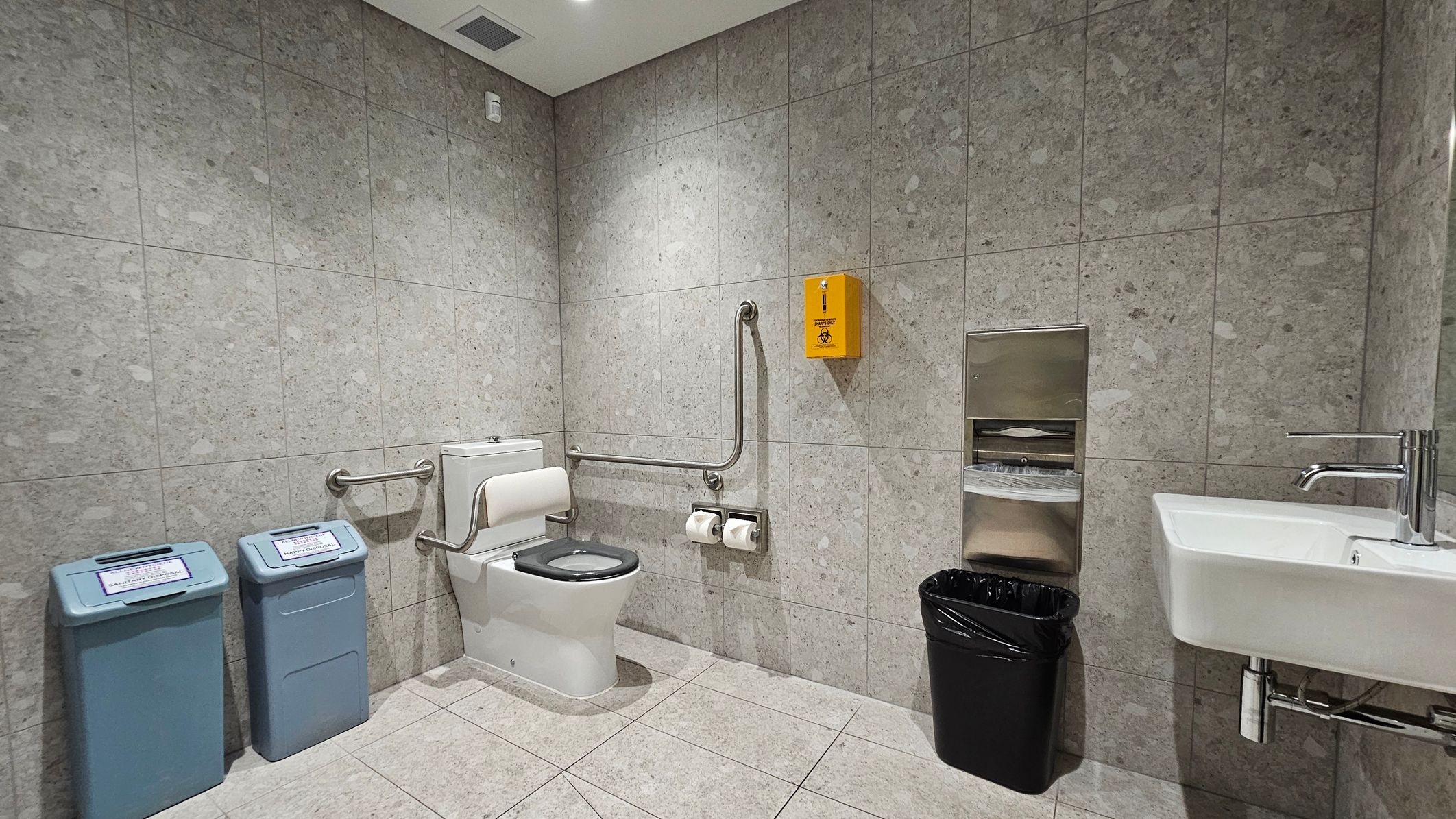 A public restroom with grey-green tiled walls and floor. On the left, there are two blue waste bins with labels. In the centre, a toilet with a black seat is equipped with support rails and two toilet paper rolls. Above the toilet, a yellow biohazard disposal box is mounted. On the right, a white sink with a silver tap is next to a black waste bin. Above the bin, a metal paper towel dispenser is attached to the wall. The ceiling is white with a vent and recessed lighting.