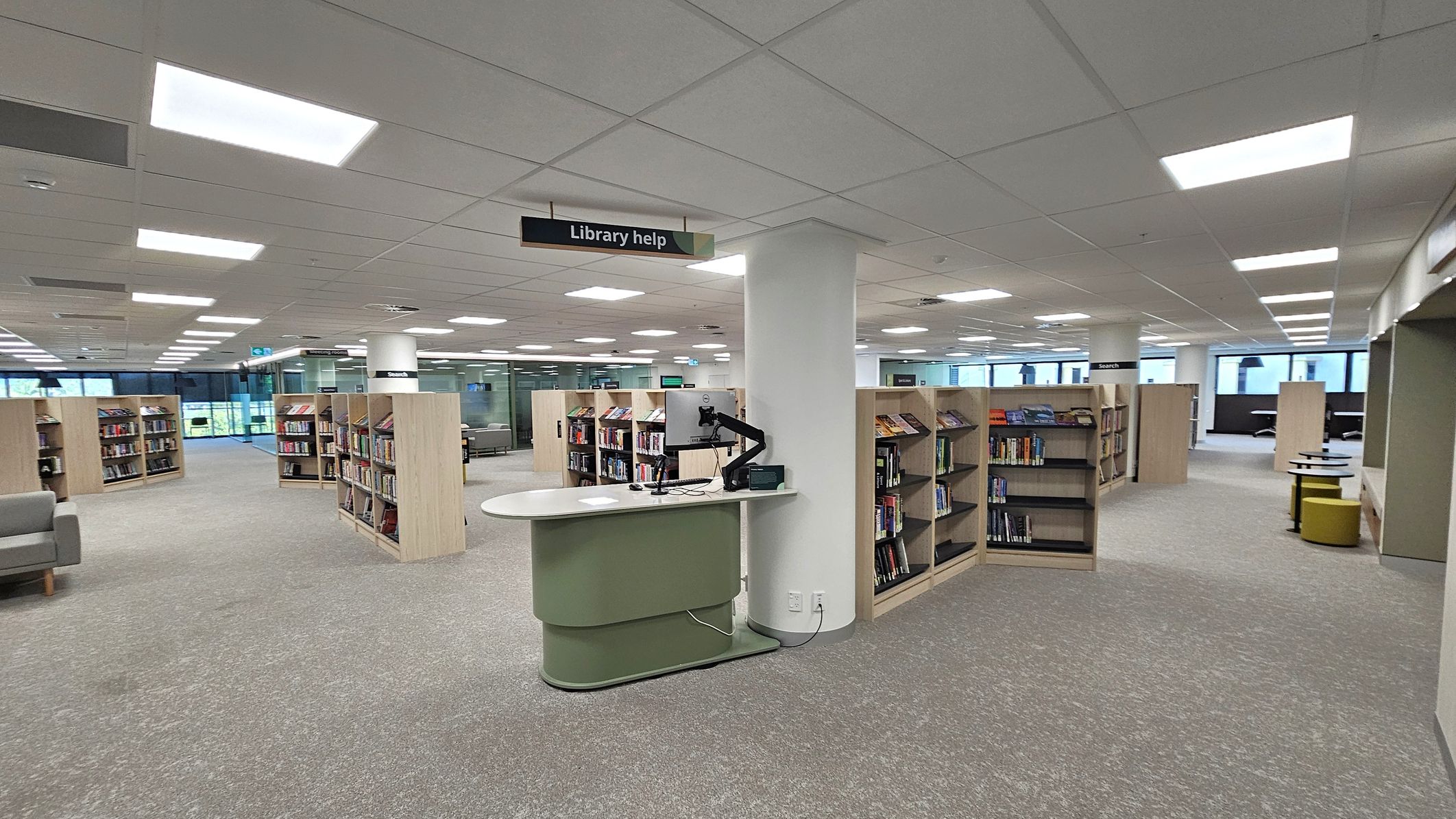 A modern library interior with a grey and green carpeted floor. In the centre, a curved help desk with a computer is positioned near a white column. Bookshelves filled with books are arranged in rows throughout the space. Overhead, a sign reads "Library help." The ceiling is fitted with rectangular fluorescent lights. Large windows in the background allow natural light to enter, and there are seating areas with grey sofas and small tables.