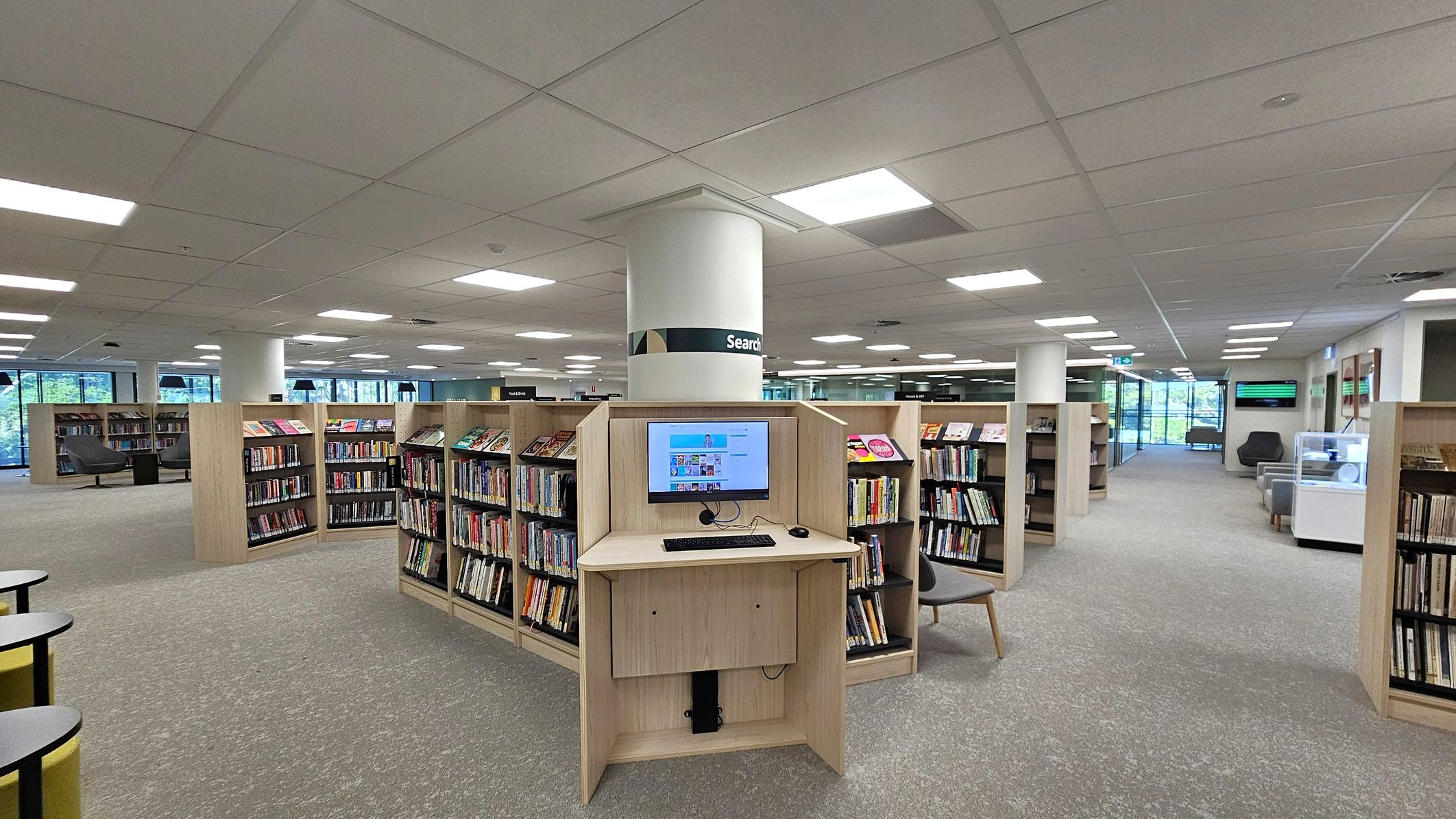 A modern library interior with a central computer station in front of a column labelled "Search". Surrounding the station are wooden bookshelves filled with books. The floor is carpeted in grey, and the ceiling has recessed lighting. Large windows on the left allow natural light to enter, and there are seating areas with grey chairs. A display case is visible on the right, and the space is open and well-lit.