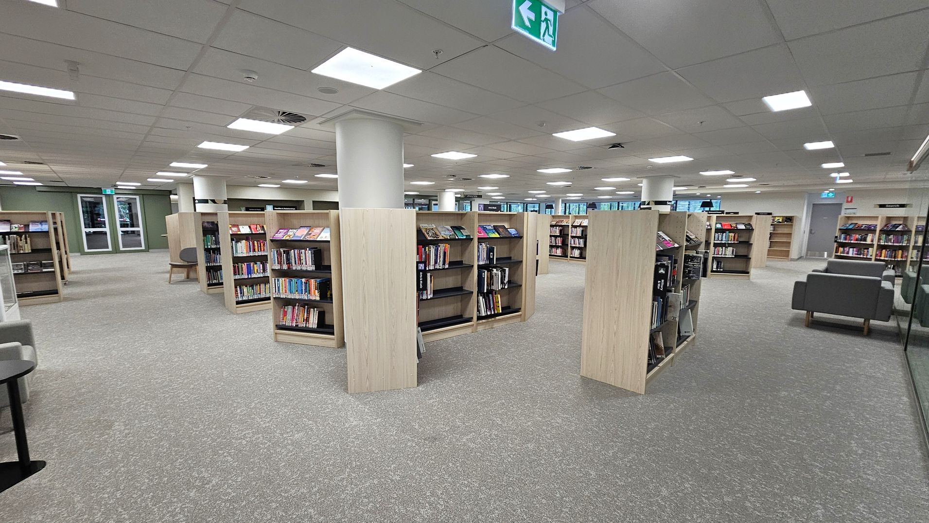 A spacious library interior with grey carpeted flooring and a grid-patterned ceiling with recessed lighting. In the centre, there are several light wood bookshelves filled with books. To the right, there are grey upholstered chairs. The background features large windows and more bookshelves. An emergency exit sign is visible on the ceiling.
