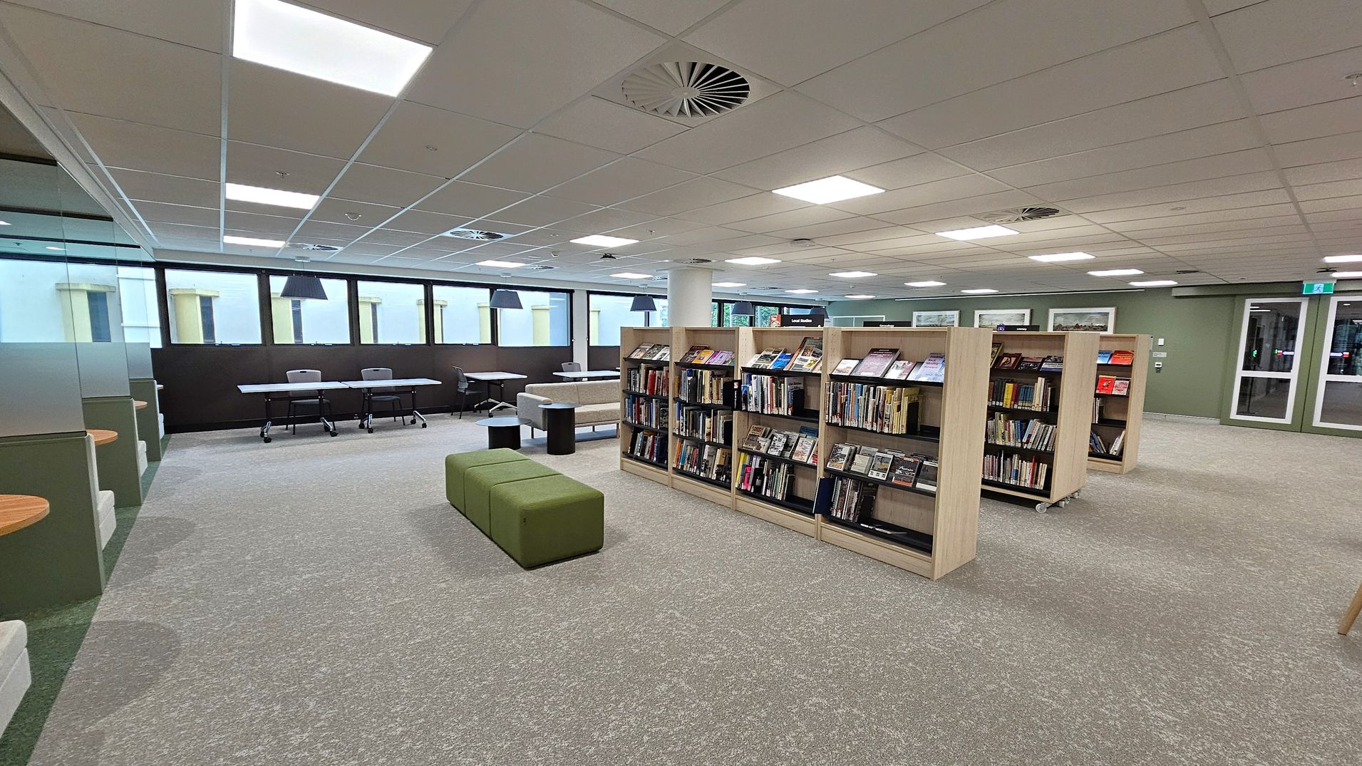 A modern library interior with a green carpeted floor. In the centre, there are wooden bookshelves filled with books and magazines. To the left, there are tables and chairs near large windows, with pendant lights hanging above. A green cushioned bench is in front of the shelves. The ceiling is fitted with square lights and air vents. In the background, there are more tables and a glass door leading to another area.