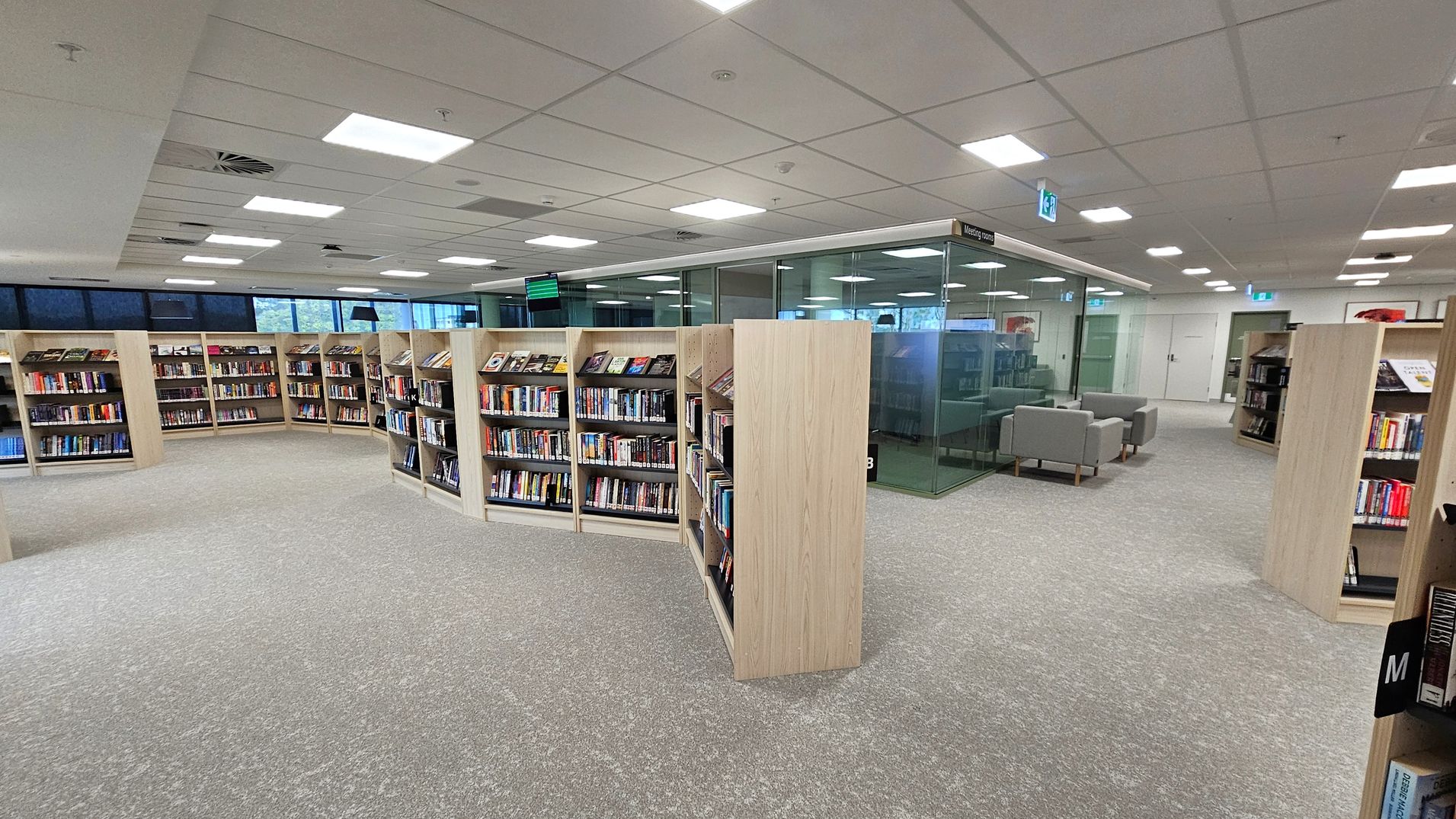 Bookshelves filled with books are arranged in a semi-circle on a grey carpeted floor. In the background, there is a glass-walled meeting space with grey sofas. The ceiling is fitted with square lights and ventilation panels. Large windows line the left side, allowing natural light to enter.