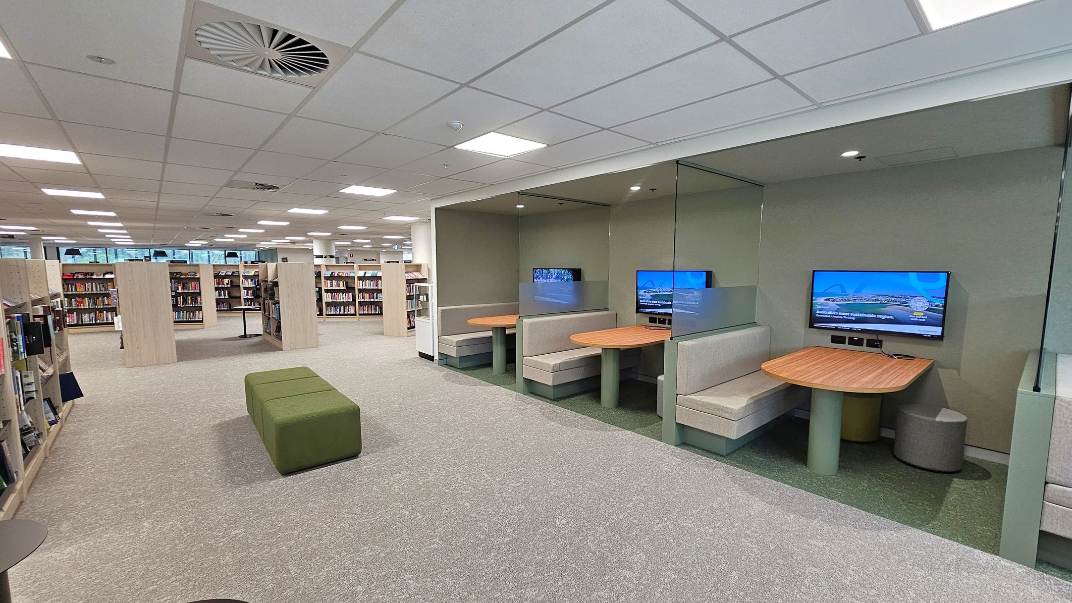 A modern library interior with a beige carpeted floor. On the right, there are two study booths with wooden tables, cushioned seating, and wall-mounted screens. The booths have green walls and are separated by glass partitions. In the centre, a green upholstered bench is placed on the floor. On the left, there are rows of bookshelves filled with books. The ceiling is white with recessed lighting and circular air vents.