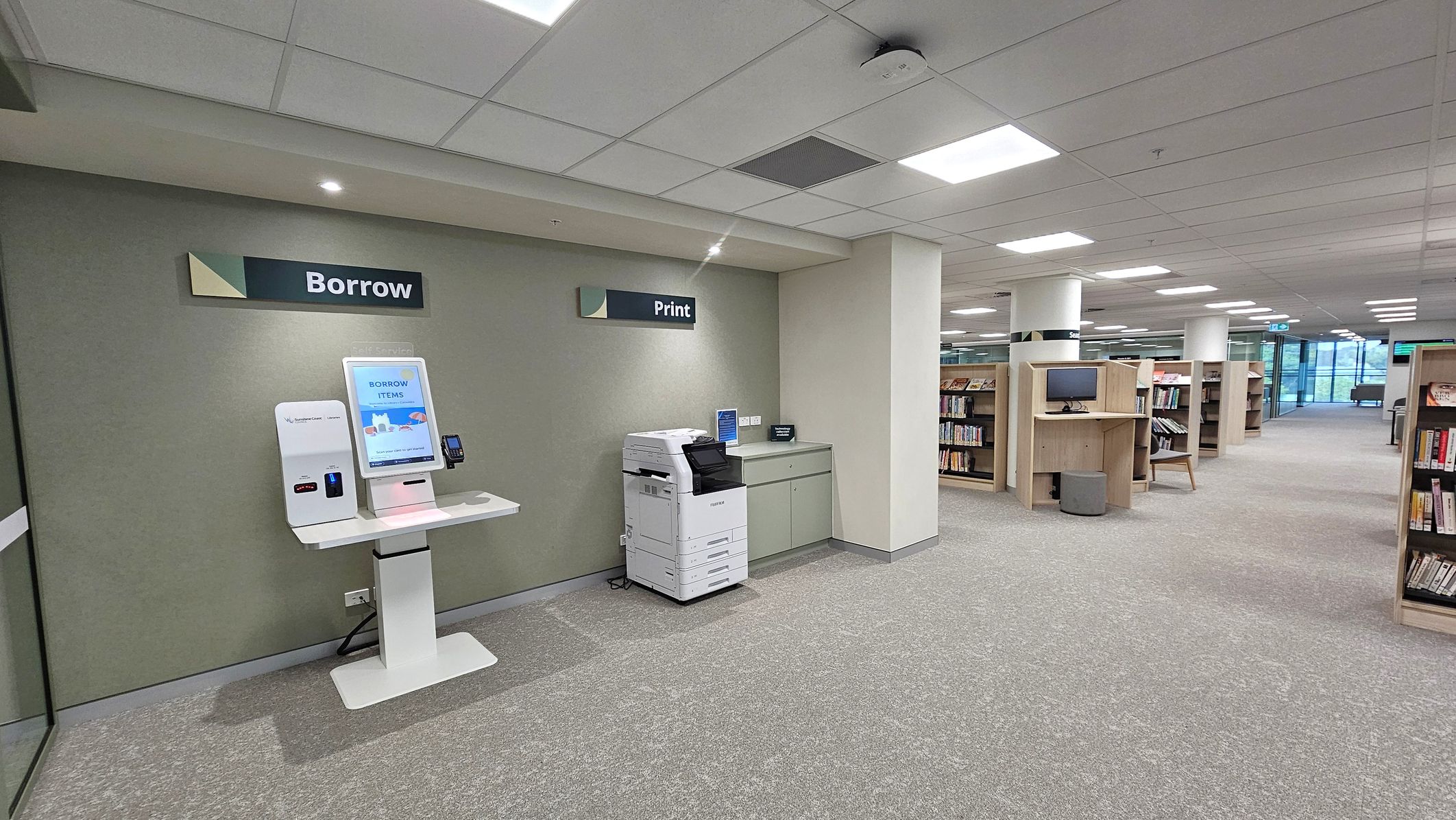 On the left, a library borrowing station with a touchscreen kiosk labelled "Borrow" and a card reader. Next to it, a printer under a "Print" sign, with a counter and power outlets. The floor is carpeted in grey. On the right, rows of wooden bookshelves and a computer station. The ceiling is white with recessed lighting. In the background, large windows provide natural light.