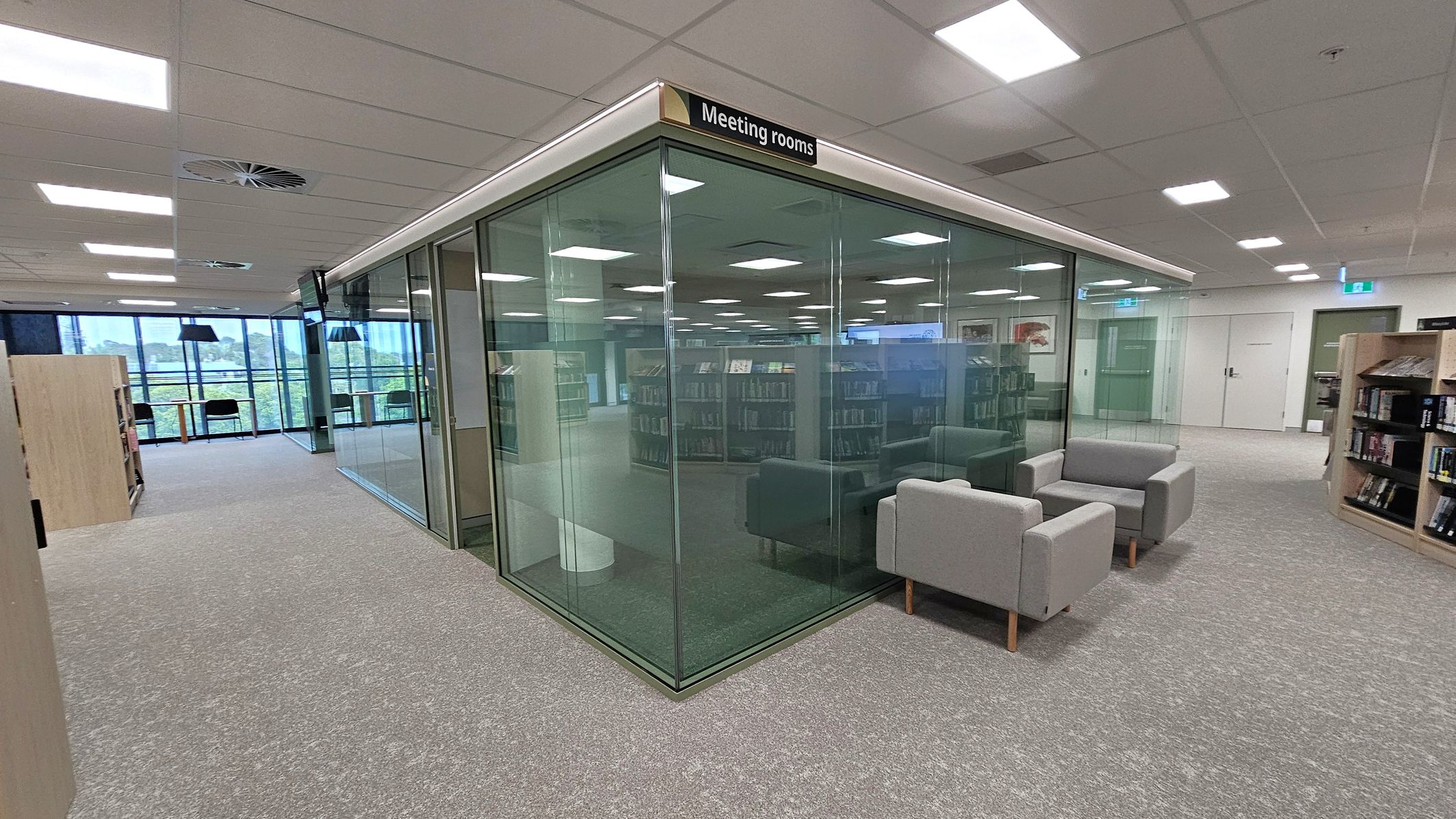 A modern library interior with a glass-walled meeting room in the centre, labelled "Meeting rooms" at the top. To the right, there are two grey armchairs. On the left, bookshelves are visible, and in the background, large windows provide natural light. The floor is carpeted in grey, and the ceiling features recessed lighting and ventilation panels.