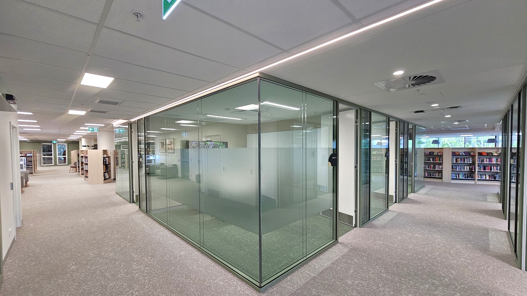 A modern library interior with a beige and grey carpeted floor. On the left, there are bookshelves and seating areas. In the centre, a glass-walled room with frosted panels is visible. On the right, more bookshelves are lined up against the wall. The ceiling is fitted with recessed lighting and air vents. An illuminated exit sign is visible above the corridor.