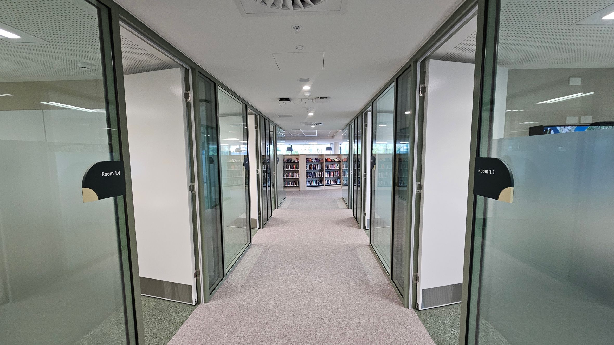 A corridor with glass-walled rooms on both sides, labelled "Room 1.4" on the left and "Room 1.1" on the right. The floor is covered with a mix of grey and pink carpet. At the end of the corridor, there are bookshelves filled with books. The ceiling is white with recessed lighting and ventilation panels.