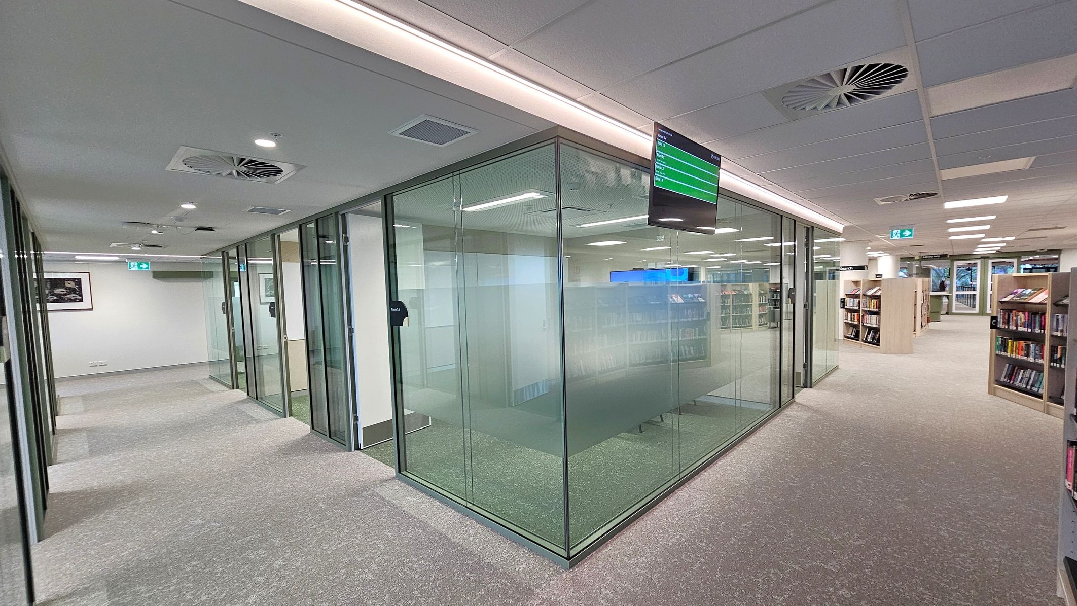 A modern library interior with glass-walled rooms on the left, featuring frosted panels for privacy. A digital screen is mounted above, displaying information. The right side has bookshelves filled with books, extending into the background. The floor is carpeted in a neutral tone, and the ceiling is fitted with recessed lighting and air vents. Emergency exit signs are visible, and framed artwork is hung on the walls.