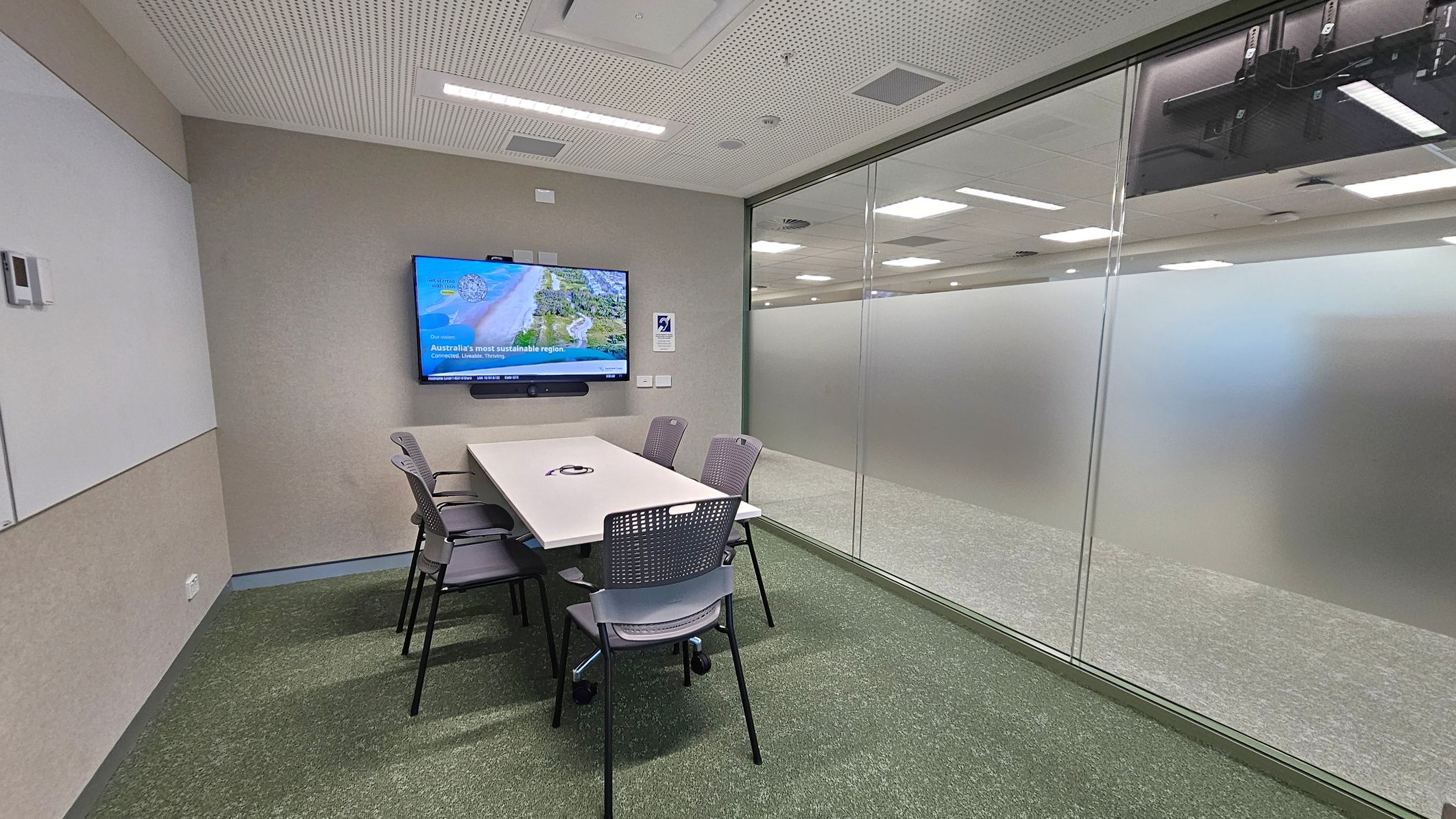 A small meeting room with a rectangular white table surrounded by six grey chairs. On the left wall, a large screen displays an image and text. The right side features a glass wall with a frosted lower section, revealing a view of an adjacent room with ceiling lights. The floor is carpeted in green, and the ceiling is white with recessed lighting and ventilation panels.