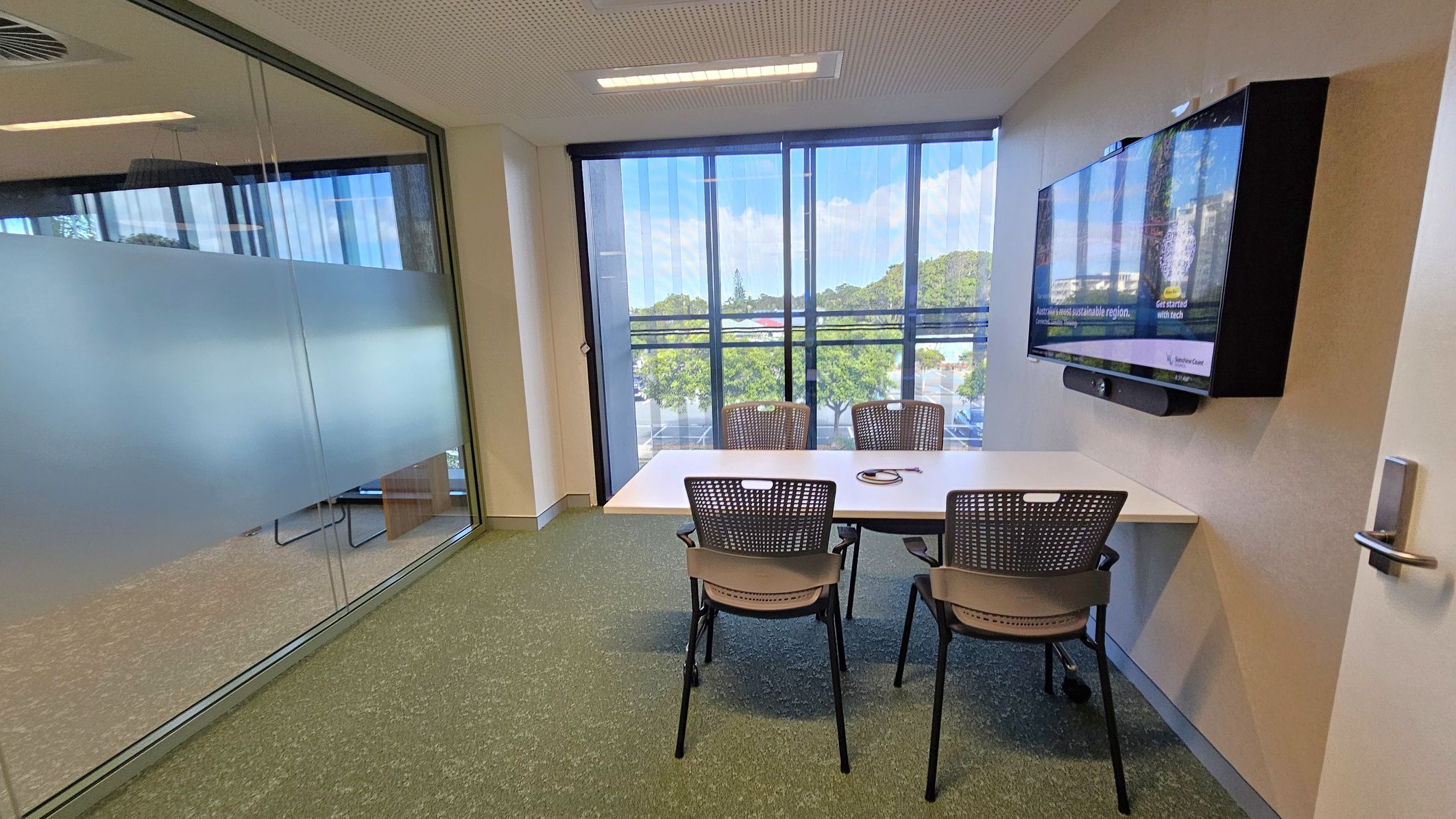 A small meeting room with a glass wall on the left, partially frosted for privacy. In the centre, a rectangular table with three chairs. On the right wall, a large flat-screen monitor is mounted. The room has a green carpet and a white ceiling with recessed lighting. The far wall features large windows with vertical blinds, offering a view of trees and a parking area outside. A door with a handle is visible on the right.