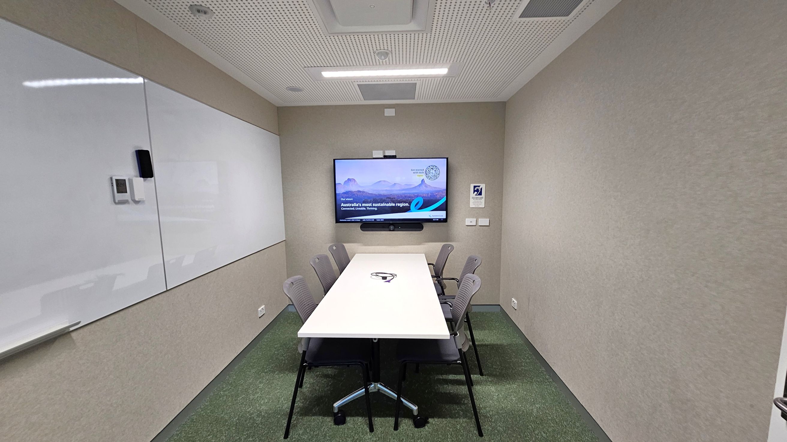 A small meeting room with a long white table in the centre surrounded by six grey chairs. On the far wall, a large screen displays a presentation. The left wall features a large whiteboard. The floor is carpeted in green, and the ceiling is white with recessed lighting and ventilation panels.