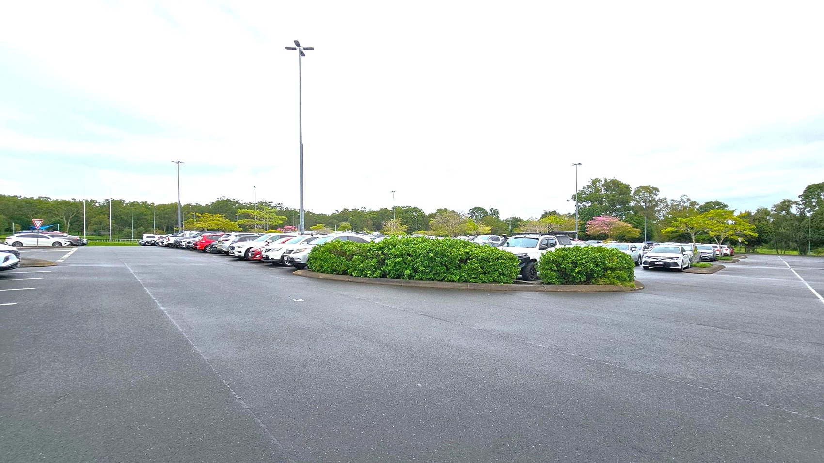 A car park with multiple vehicles parked in rows. In the centre, there is a landscaped area with green bushes. The ground is paved with asphalt, and tall streetlights are visible. In the background, there are trees and greenery, providing a natural border to the parking area. A car park with multiple vehicles parked in rows. In the centre, there is a landscaped area with green bushes. The ground is paved with asphalt, and tall streetlights are visible. In the background, there are trees and greenery, providing a natural border to the parking area.