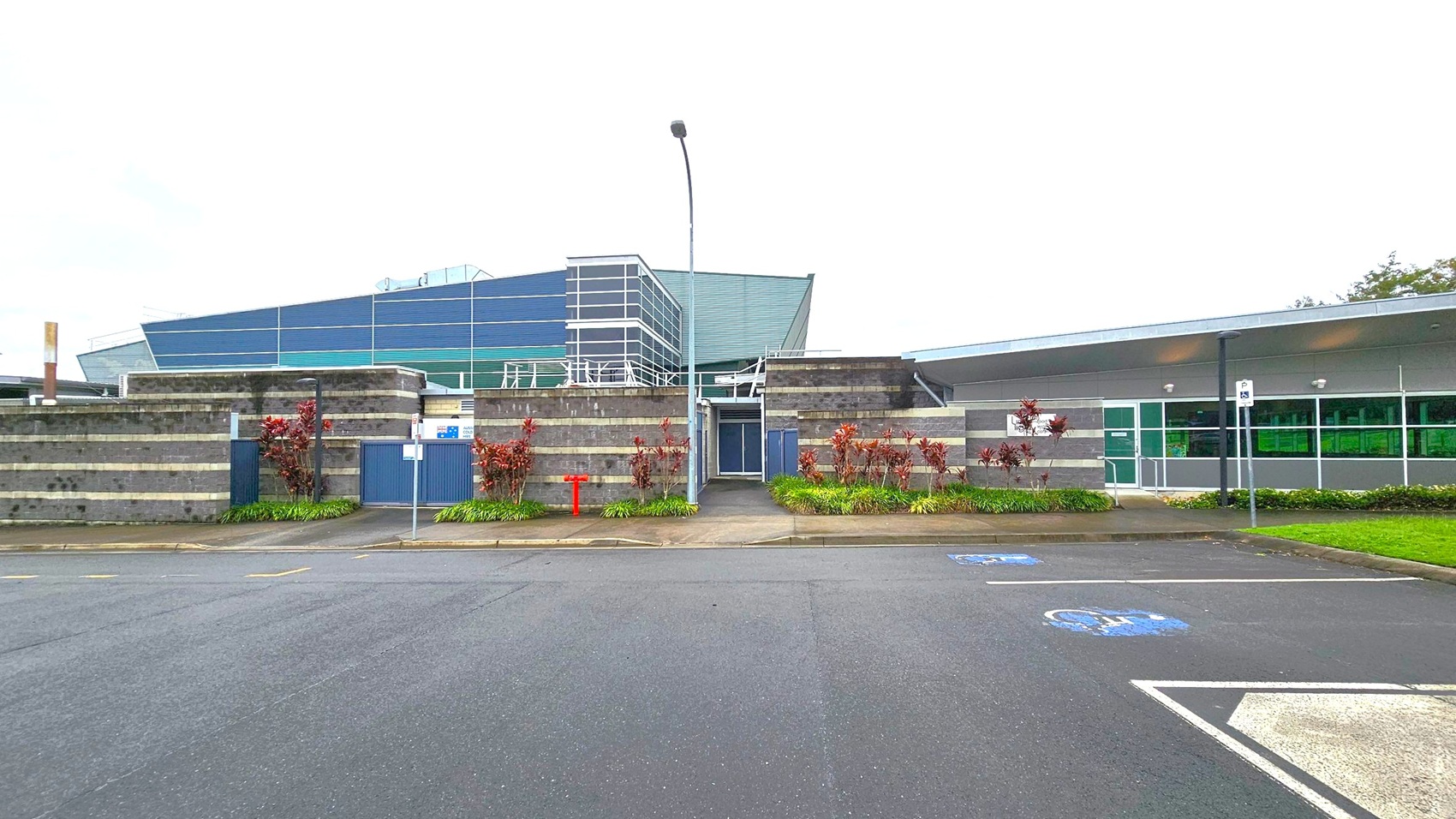 A modern building with a mix of grey and green panels. The left side features a structure with horizontal stripes and a central entrance, flanked by small plants. The right side has large windows and a flat roof. In the foreground, there is a paved area with marked parking spaces, including a disabled parking spot. A tall streetlamp stands in front of the entrance. The surrounding area is landscaped with grass and shrubs. A modern building with a mix of grey and green panels. The left side features a structure with horizontal stripes and a central entrance, flanked by small plants. The right side has large windows and a flat roof. In the foreground, there is a paved area with marked parking spaces, including a disabled parking spot. A tall streetlamp stands in front of the entrance. The surrounding area is landscaped with grass and shrubs.