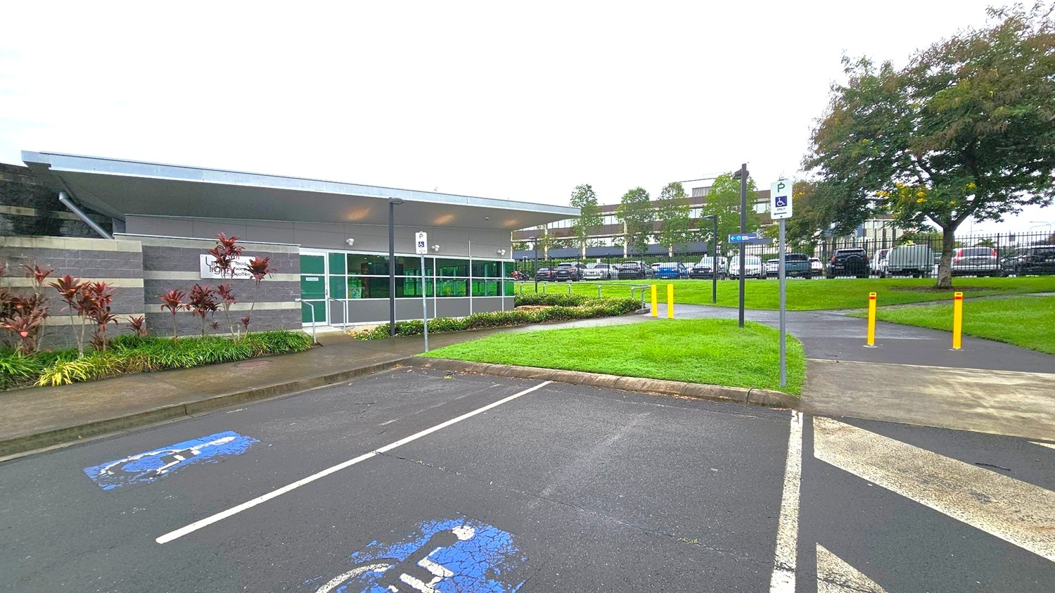 A modern building with a flat roof is on the left, featuring grey and green panels and a sign. In front, there are landscaped plants and a pathway. The foreground shows a car park with a marked disabled parking space. On the right, a grassy area with yellow bollards and a paved path leads to a fence. Beyond the fence, there are parked cars and trees. The sky is overcast. A modern building with a flat roof is on the left, featuring grey and green panels and a sign. In front, there are landscaped plants and a pathway. The foreground shows a car park with a marked disabled parking space. On the right, a grassy area with yellow bollards and a paved path leads to a fence. Beyond the fence, there are parked cars and trees. The sky is overcast.