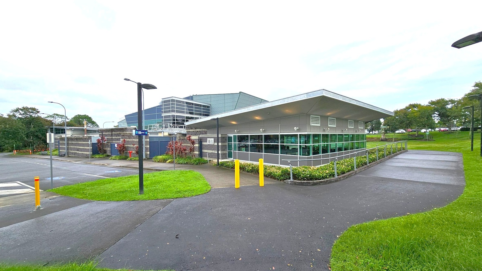 A modern building with large glass windows and a sloped roof is situated on the right. It is surrounded by a well-maintained lawn and a paved path with yellow bollards. On the left, there is a parking area with a few street lamps and signs. In the background, trees and more greenery are visible, along with a larger building with a glass facade. The sky is overcast, and the ground is wet, suggesting recent rain. A modern building with large glass windows and a sloped roof is situated on the right. It is surrounded by a well-maintained lawn and a paved path with yellow bollards. On the left, there is a parking area with a few street lamps and signs. In the background, trees and more greenery are visible, along with a larger building with a glass facade. The sky is overcast, and the ground is wet, suggesting recent rain.
