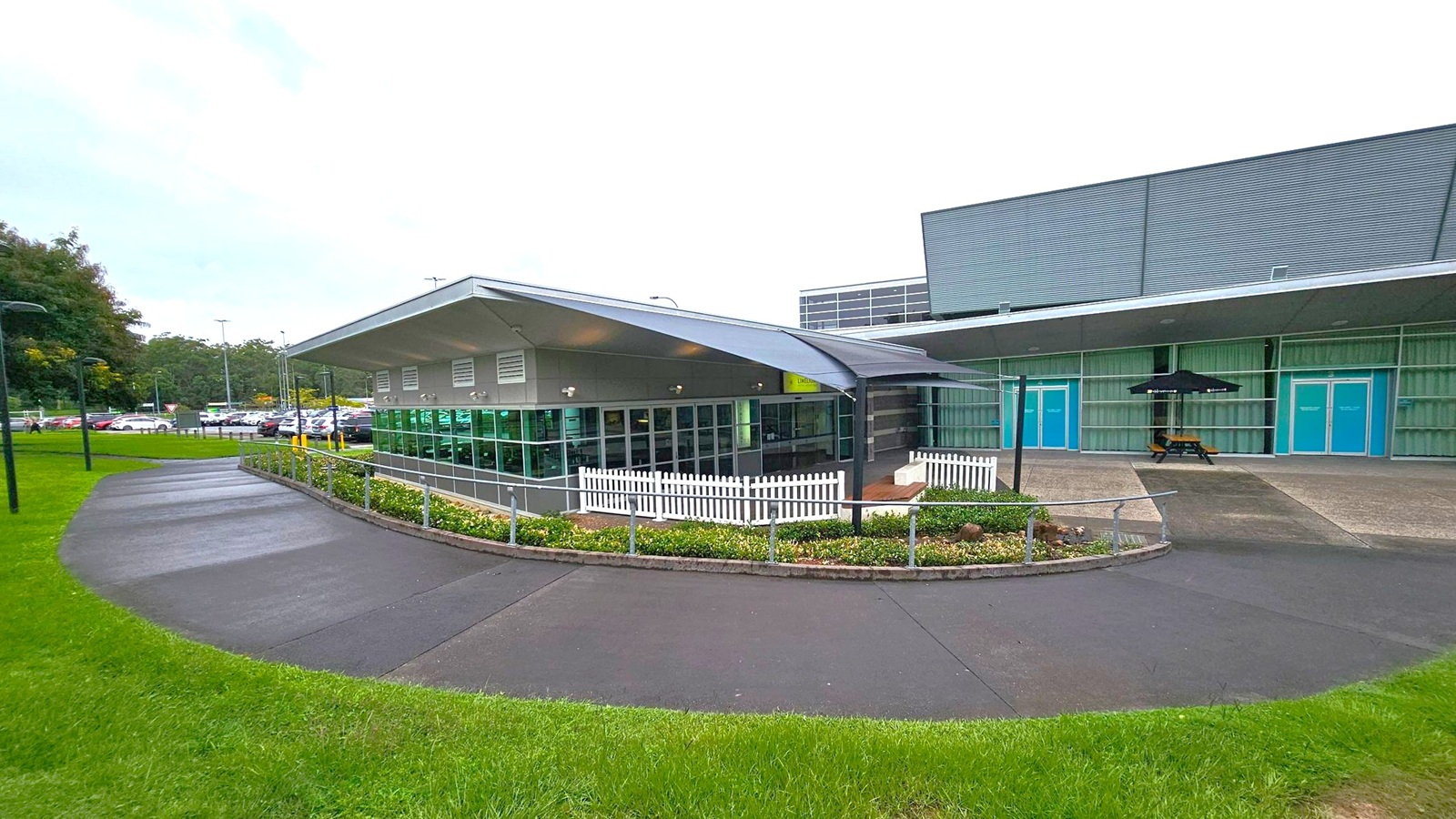 A modern building with a sloped roof and large glass windows on the left. A white picket fence surrounds a small garden area. The right side features a larger structure with blue doors and a covered area with a picnic table and umbrella. The foreground has a curved pathway bordered by green grass, leading to a car park visible in the background. A modern building with a sloped roof and large glass windows on the left. A white picket fence surrounds a small garden area. The right side features a larger structure with blue doors and a covered area with a picnic table and umbrella. The foreground has a curved pathway bordered by green grass, leading to a car park visible in the background.