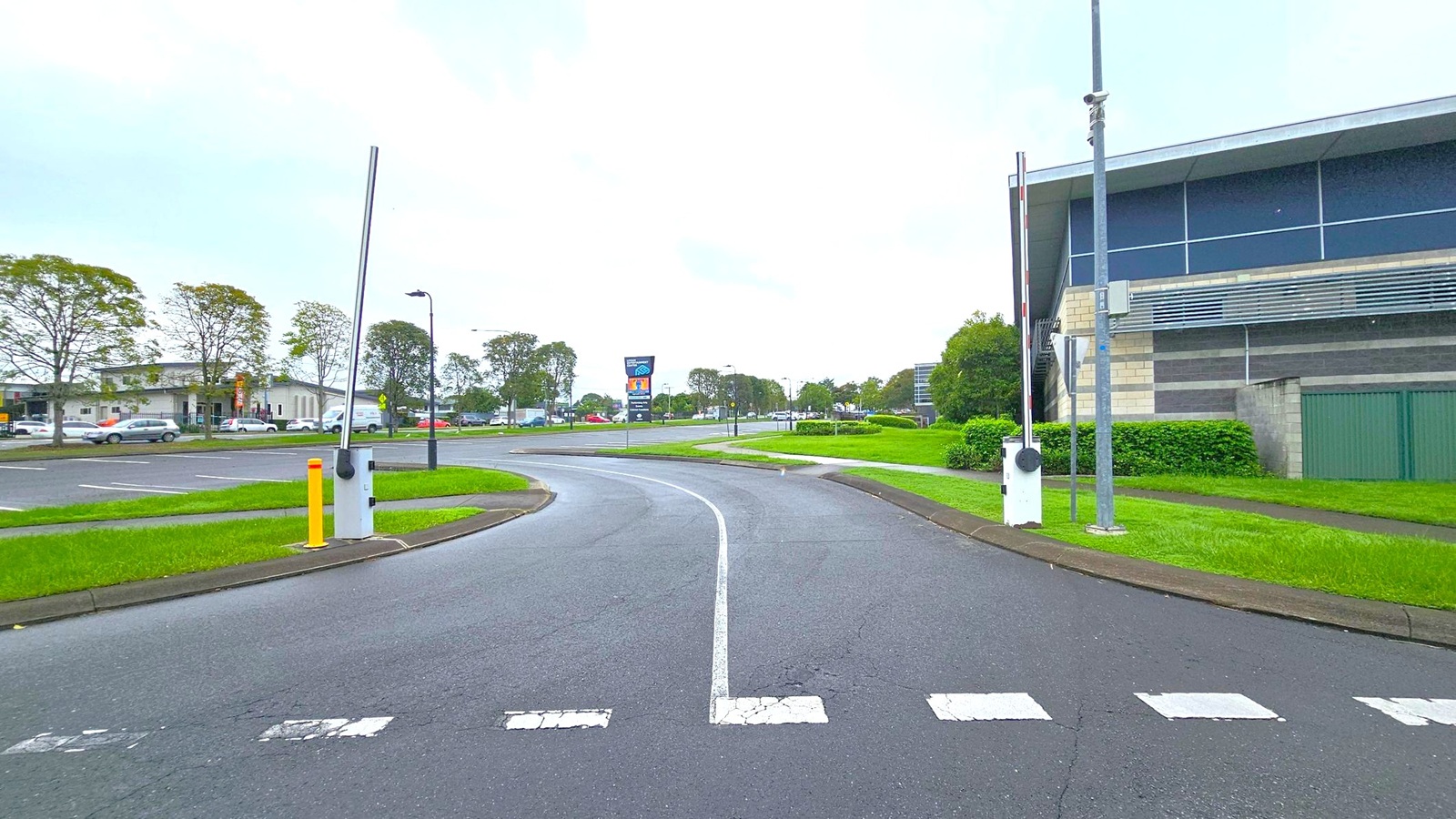 A road with a central lane curves into the distance, flanked by grassy verges. On the right, a modern building with large windows and a green fence is visible. A tall pole with a barrier arm stands nearby. On the left, a parking area with several cars and a row of trees can be seen. In the background, a large sign and more trees line the road. The sky is overcast, creating a muted atmosphere. A road with a central lane curves into the distance, flanked by grassy verges. On the right, a modern building with large windows and a green fence is visible. A tall pole with a barrier arm stands nearby. On the left, a parking area with several cars and a row of trees can be seen. In the background, a large sign and more trees line the road. The sky is overcast, creating a muted atmosphere.