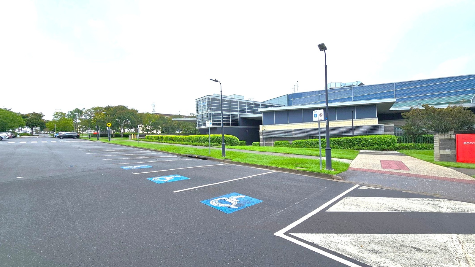 A parking area with marked spaces, including disabled parking spots with blue symbols on the asphalt. On the right, a modern building with large glass windows and a flat roof is visible, surrounded by neatly trimmed hedges. A pathway with a red and beige pattern leads to the building. Street lamps line the path, and a red sign is partially visible on the right. Trees and parked cars are on the left, with a road running alongside. A parking area with marked spaces, including disabled parking spots with blue symbols on the asphalt. On the right, a modern building with large glass windows and a flat roof is visible, surrounded by neatly trimmed hedges. A pathway with a red and beige pattern leads to the building. Street lamps line the path, and a red sign is partially visible on the right. Trees and parked cars are on the left, with a road running alongside.