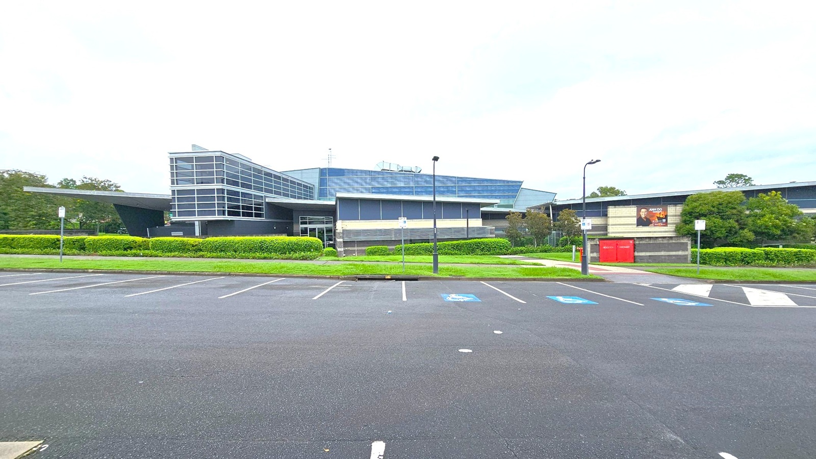 A modern building with a glass facade and angular design is in the centre, surrounded by well-maintained hedges and greenery. To the right, a billboard is mounted on a wall next to red utility boxes. The foreground features an empty car park with marked spaces, including accessible parking spots. Street lamps line the pathway leading to the building. A modern building with a glass facade and angular design is in the centre, surrounded by well-maintained hedges and greenery. To the right, a billboard is mounted on a wall next to red utility boxes. The foreground features an empty car park with marked spaces, including accessible parking spots. Street lamps line the pathway leading to the building.
