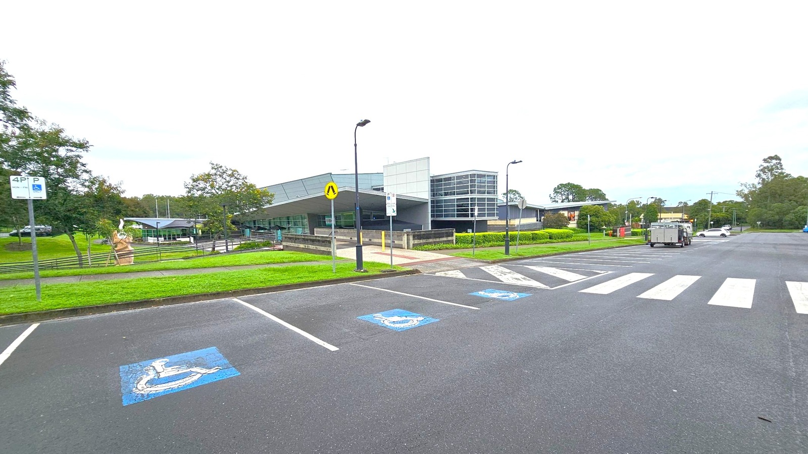 A modern building with large glass windows is in the centre, surrounded by green lawns and trees. On the left, a sculpture is visible near a pathway. The foreground features a car park with marked disabled parking spaces and a zebra crossing. A few street lamps are scattered around, and a white car is parked on the right side. The area is bordered by more greenery and trees in the background. A modern building with large glass windows is in the centre, surrounded by green lawns and trees. On the left, a sculpture is visible near a pathway. The foreground features a car park with marked disabled parking spaces and a zebra crossing. A few street lamps are scattered around, and a white car is parked on the right side. The area is bordered by more greenery and trees in the background.