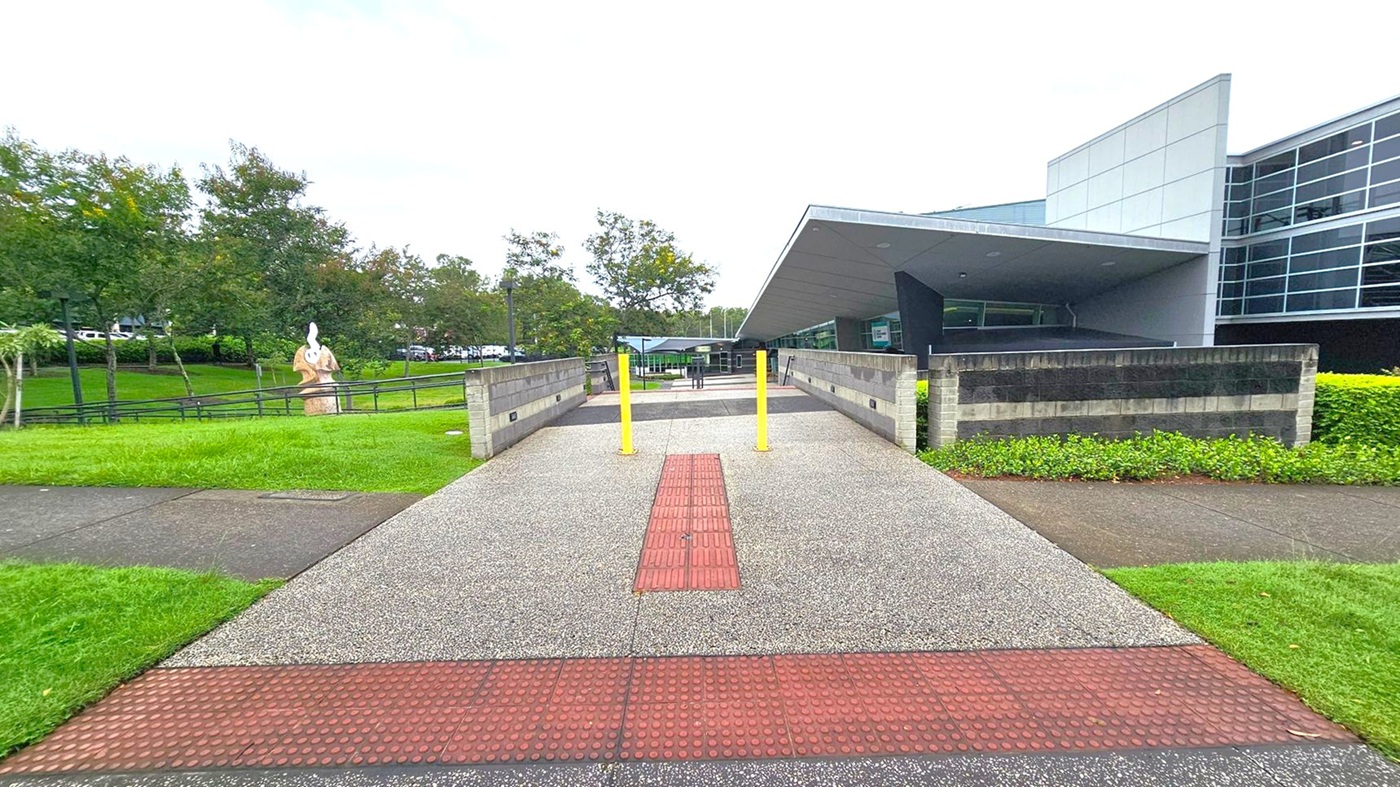 A pathway with a textured red strip runs through the centre, flanked by two yellow bollards. On the right, a modern building with large glass windows and a slanted roof is visible. To the left, a grassy area with trees and a sculpture can be seen. The pathway is bordered by low brick walls and greenery. The sky is overcast. A pathway with a textured red strip runs through the centre, flanked by two yellow bollards. On the right, a modern building with large glass windows and a slanted roof is visible. To the left, a grassy area with trees and a sculpture can be seen. The pathway is bordered by low brick walls and greenery. The sky is overcast.
