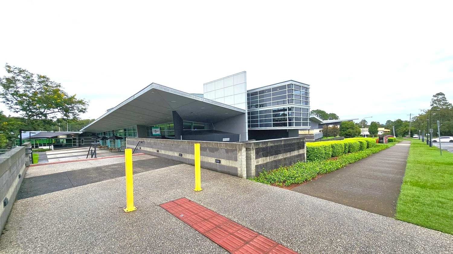 A modern building with a slanted roof and large glass windows is in the centre. The foreground features a grey, pebbled pathway with a red tactile strip and two yellow bollards. On the right, a neatly trimmed hedge lines a concrete footpath, with a grassy area beside it. To the left, a tree and more pathways are visible. The background includes more trees and a few parked cars. A modern building with a slanted roof and large glass windows is in the centre. The foreground features a grey, pebbled pathway with a red tactile strip and two yellow bollards. On the right, a neatly trimmed hedge lines a concrete footpath, with a grassy area beside it. To the left, a tree and more pathways are visible. The background includes more trees and a few parked cars.