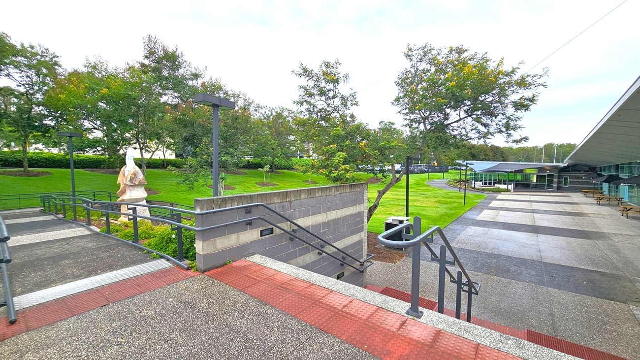 A paved outdoor area with a series of ramps and stairs in the foreground, featuring metal railings. On the left, a stone sculpture stands amidst green grass and trees. The background has a modern building with a sloped roof and glass windows. Several picnic tables are placed along the right side, near the building. The area is well-maintained, with neatly trimmed grass and scattered trees. A paved outdoor area with a series of ramps and stairs in the foreground, featuring metal railings. On the left, a stone sculpture stands amidst green grass and trees. The background has a modern building with a sloped roof and glass windows. Several picnic tables are placed along the right side, near the building. The area is well-maintained, with neatly trimmed grass and scattered trees.