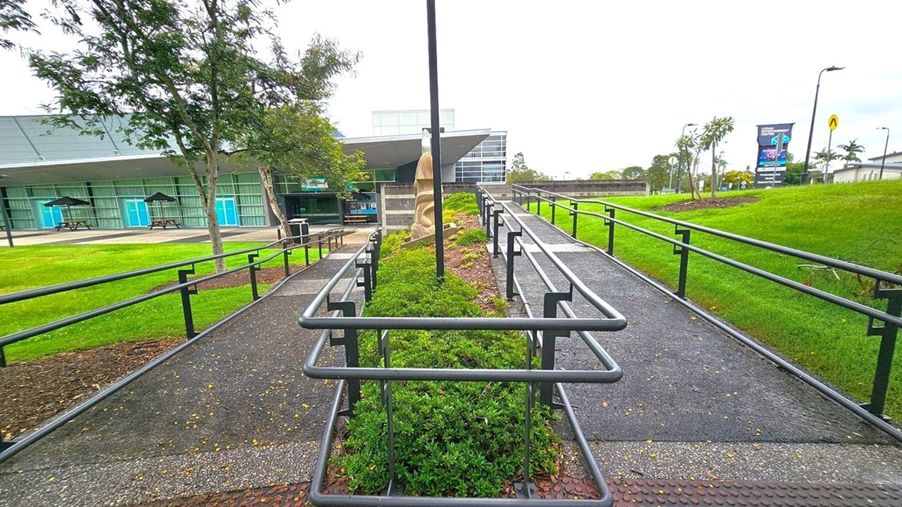 A pathway with metal railings runs through the centre, flanked by green shrubs. On the left, a building with large windows and blue doors is visible, with picnic tables and umbrellas outside. On the right, a grassy area with a few small trees and a sign in the distance. A sculpture is positioned in the middle of the pathway. The sky is overcast, and the ground is paved with small stones. A pathway with metal railings runs through the centre, flanked by green shrubs. On the left, a building with large windows and blue doors is visible, with picnic tables and umbrellas outside. On the right, a grassy area with a few small trees and a sign in the distance. A sculpture is positioned in the middle of the pathway. The sky is overcast, and the ground is paved with small stones.