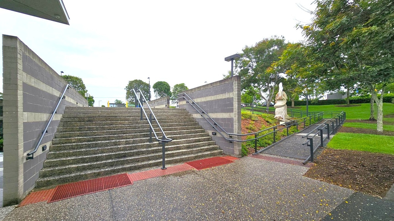 Wide stone steps with metal railings in the centre, flanked by grey brick walls. To the right, a sloped path with railings leads up, bordered by grass and trees. A statue stands on the right side of the path amidst greenery. The ground is speckled with small leaves or debris. In the background, more trees and a few lamp posts are visible. Wide stone steps with metal railings in the centre, flanked by grey brick walls. To the right, a sloped path with railings leads up, bordered by grass and trees. A statue stands on the right side of the path amidst greenery. The ground is speckled with small leaves or debris. In the background, more trees and a few lamp posts are visible.