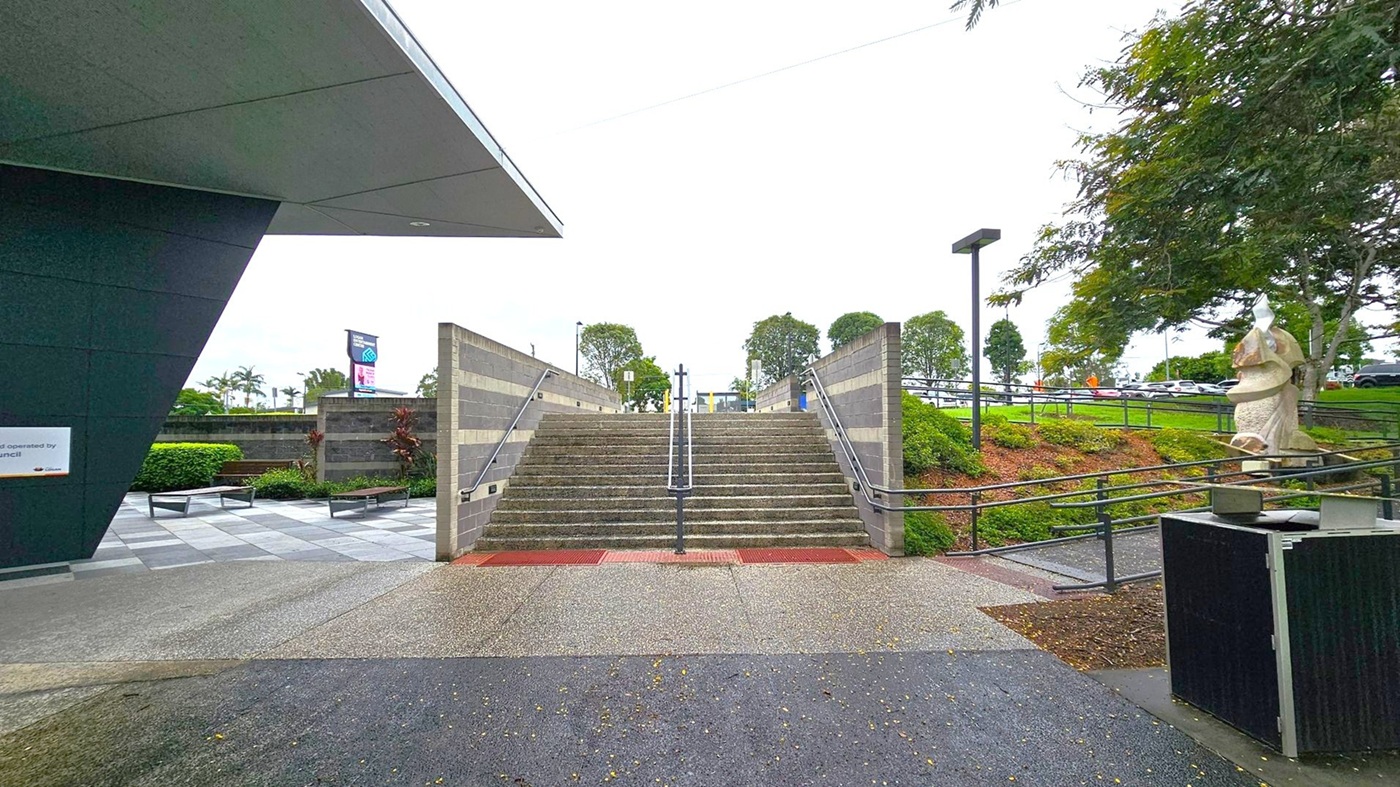 A set of wide stone steps with metal railings in the centre leads upwards, flanked by grey brick walls. On the left, a modern building with a dark overhanging roof and a sign is visible. To the right, a large sculpture stands near a tree, with a grassy area and a path leading to a car park in the background. The ground is a mix of concrete and grass, and there are benches and small shrubs on the left side. A set of wide stone steps with metal railings in the centre leads upwards, flanked by grey brick walls. On the left, a modern building with a dark overhanging roof and a sign is visible. To the right, a large sculpture stands near a tree, with a grassy area and a path leading to a car park in the background. The ground is a mix of concrete and grass, and there are benches and small shrubs on the left side.