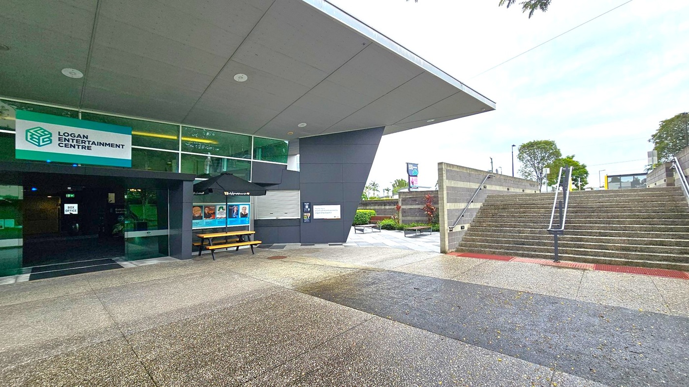 Entrance to a modern building with a sign reading "Logan Entertainment Centre" on the left. The entrance features glass doors and a box office. A bench and posters are under a black umbrella. On the right, a wide set of concrete stairs with metal railings leads upwards, flanked by grey walls. The ground is a mix of textured concrete and green patches. Trees and shrubs are visible in the background. The ceiling is flat and grey. Entrance to a modern building with a sign reading "Logan Entertainment Centre" on the left. The entrance features glass doors and a box office. A bench and posters are under a black umbrella. On the right, a wide set of concrete stairs with metal railings leads upwards, flanked by grey walls. The ground is a mix of textured concrete and green patches. Trees and shrubs are visible in the background. The ceiling is flat and grey.