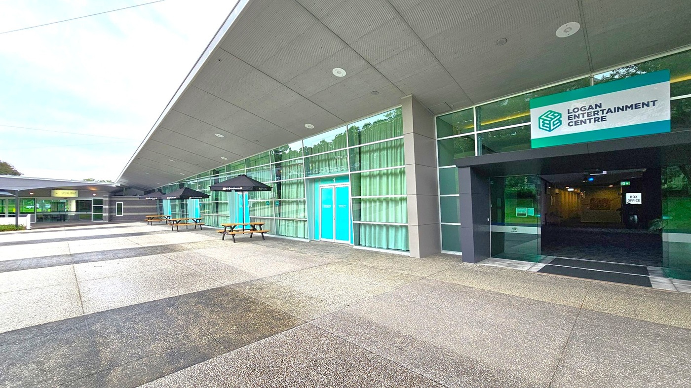 A modern building with a grey, angular roof and large glass windows. On the right, a sign reads "Logan Entertainment Centre" above a dark entrance with a "Box Office" sign inside. In the centre, blue double doors are flanked by glass panels with curtains. On the left, outdoor seating with wooden picnic tables and black umbrellas. The ground is paved with grey and speckled tiles. A modern building with a grey, angular roof and large glass windows. On the right, a sign reads "Logan Entertainment Centre" above a dark entrance with a "Box Office" sign inside. In the centre, blue double doors are flanked by glass panels with curtains. On the left, outdoor seating with wooden picnic tables and black umbrellas. The ground is paved with grey and speckled tiles.