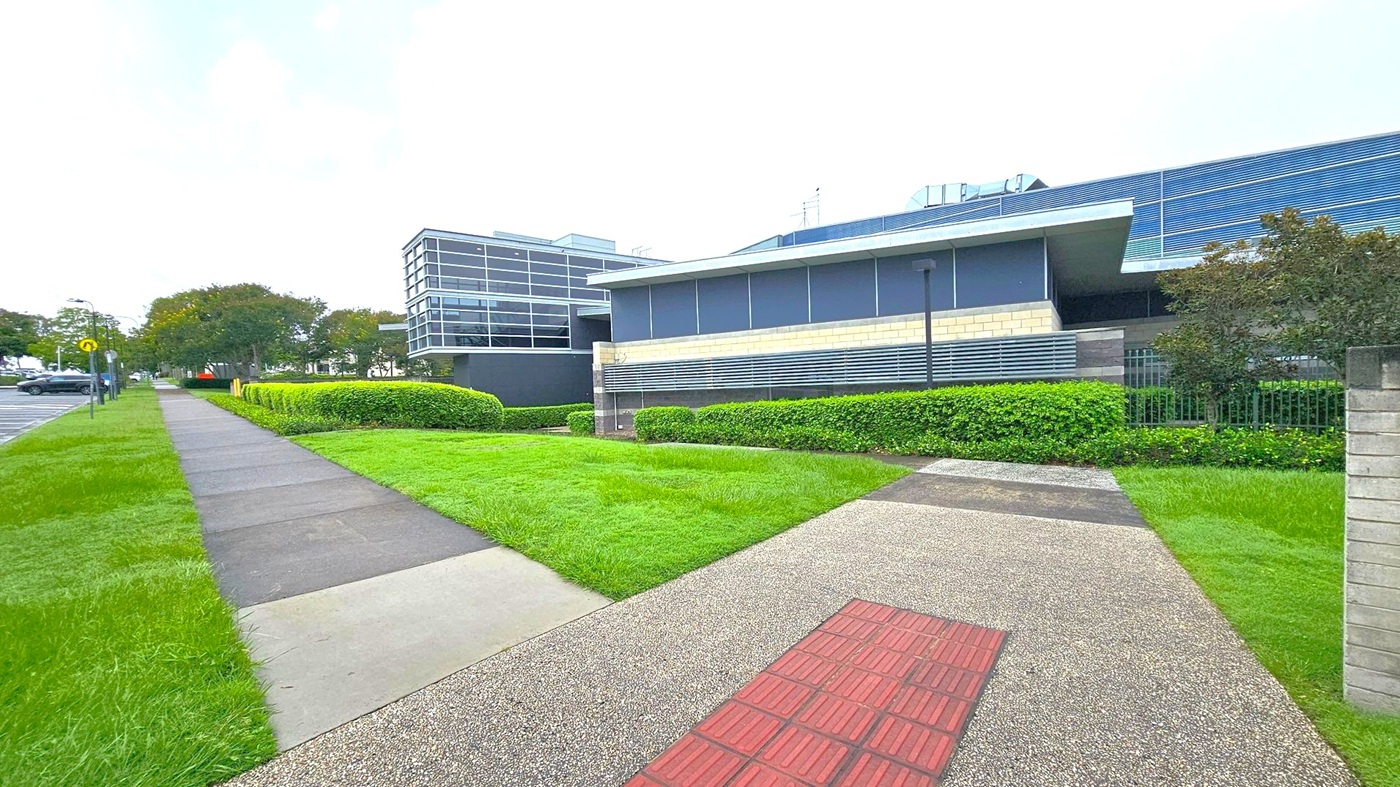 A modern building with a sleek design is on the right, featuring large windows and horizontal metal slats. In front, there's a well-maintained lawn bordered by neatly trimmed hedges. A paved path with a textured red section runs alongside the building. On the left, a concrete footpath is bordered by grass, leading towards a parking area with several cars and trees in the background. The scene is urban and orderly. A modern building with a sleek design is on the right, featuring large windows and horizontal metal slats. In front, there's a well-maintained lawn bordered by neatly trimmed hedges. A paved path with a textured red section runs alongside the building. On the left, a concrete footpath is bordered by grass, leading towards a parking area with several cars and trees in the background. The scene is urban and orderly.