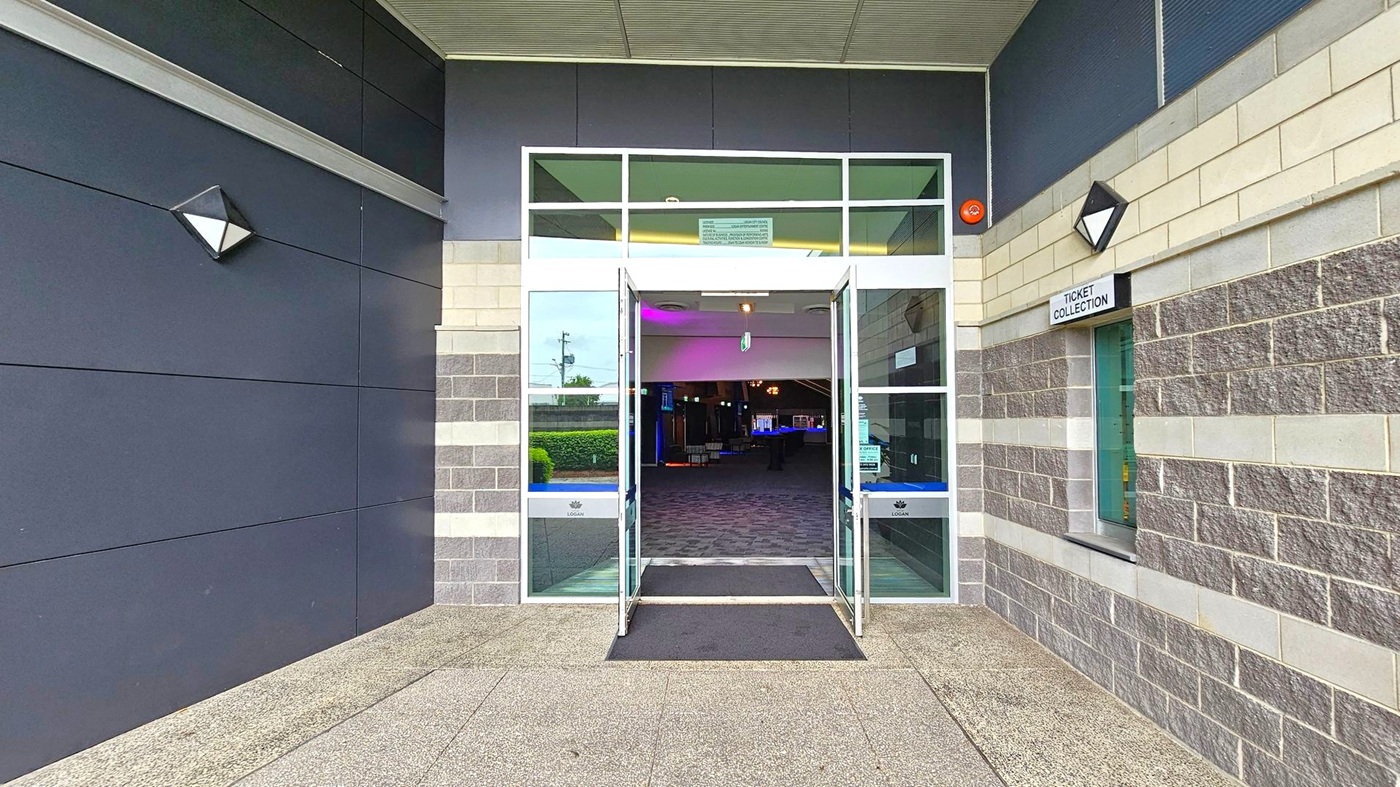 Entrance to a building with double glass doors open, leading to a dimly lit interior. The exterior walls are a mix of dark panels on the left and grey brick on the right. A sign above a window on the right reads "Ticket Collection". The floor is a speckled concrete surface, and there is a black mat in front of the doors. The ceiling is light-coloured with a grid pattern. Triangular wall lights are mounted on either side of the entrance. Entrance to a building with double glass doors open, leading to a dimly lit interior. The exterior walls are a mix of dark panels on the left and grey brick on the right. A sign above a window on the right reads "Ticket Collection". The floor is a speckled concrete surface, and there is a black mat in front of the doors. The ceiling is light-coloured with a grid pattern. Triangular wall lights are mounted on either side of the entrance.