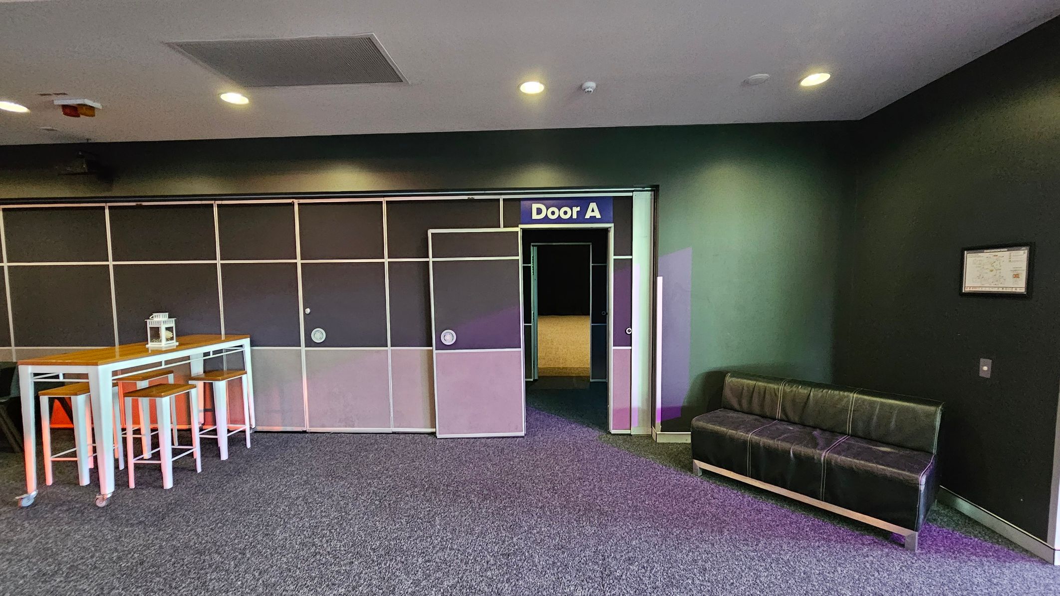 On the left, there is a tall wooden table with white metal legs, accompanied by three matching stools. A small lantern sits on the table. In the centre, a door labelled "Door A" is slightly open, revealing a dimly lit interior. On the right, a black leather sofa is positioned against a dark wall, with a framed item hanging above it. The floor is carpeted in grey, and the ceiling is fitted with recessed lights and a ventilation panel. On the left, there is a tall wooden table with white metal legs, accompanied by three matching stools. A small lantern sits on the table. In the centre, a door labelled "Door A" is slightly open, revealing a dimly lit interior. On the right, a black leather sofa is positioned against a dark wall, with a framed item hanging above it. The floor is carpeted in grey, and the ceiling is fitted with recessed lights and a ventilation panel.