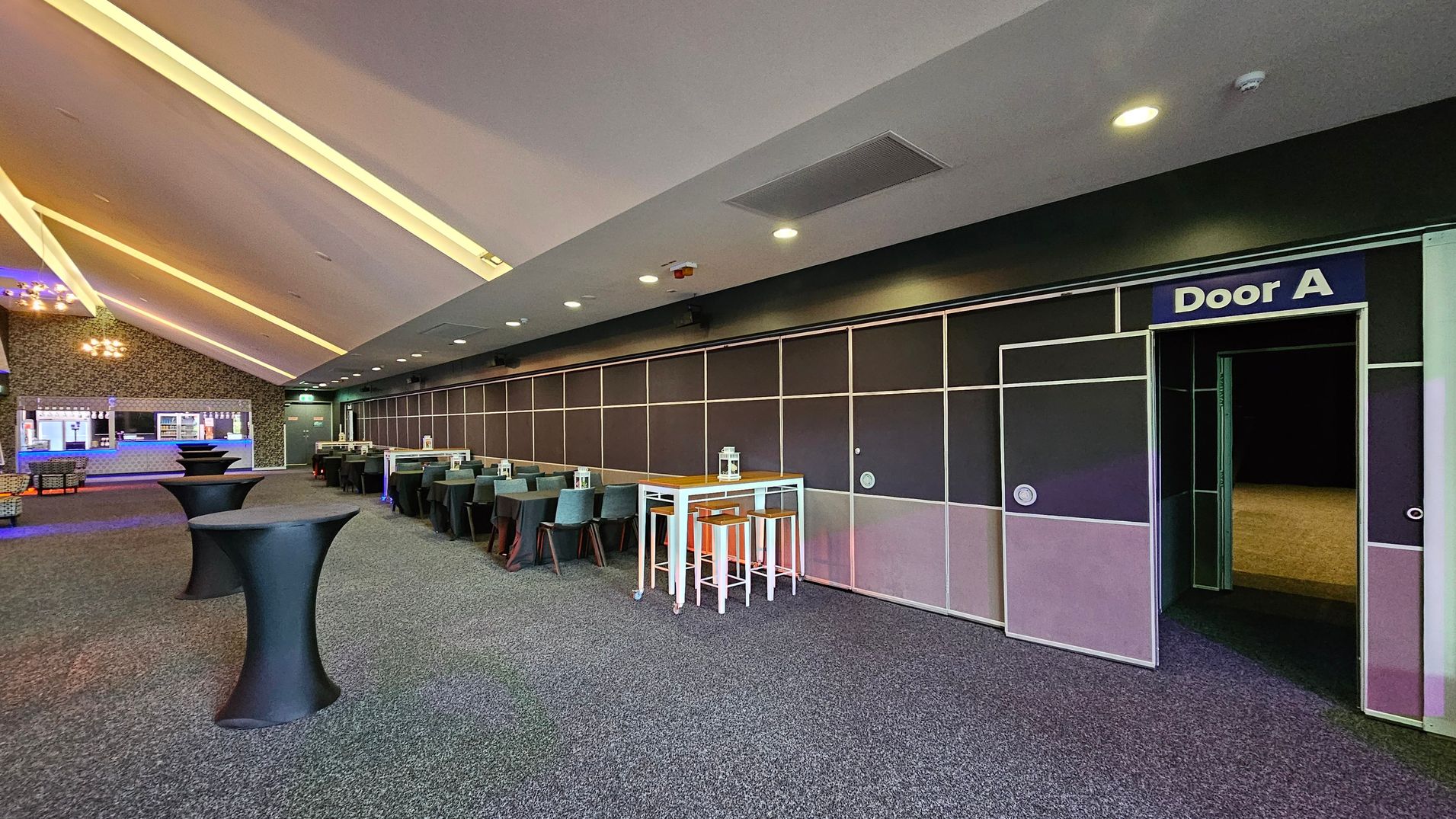 A spacious room with a grey carpeted floor and a ceiling featuring recessed lighting. On the right, a wall with a door labelled "Door A" leads to another area. In the centre, there are high tables with black covers and a white table with stools. On the left, a bar area with a counter and shelves is visible. The room has a modern design with a mix of dark and light tones. A spacious room with a grey carpeted floor and a ceiling featuring recessed lighting. On the right, a wall with a door labelled "Door A" leads to another area. In the centre, there are high tables with black covers and a white table with stools. On the left, a bar area with a counter and shelves is visible. The room has a modern design with a mix of dark and light tones.