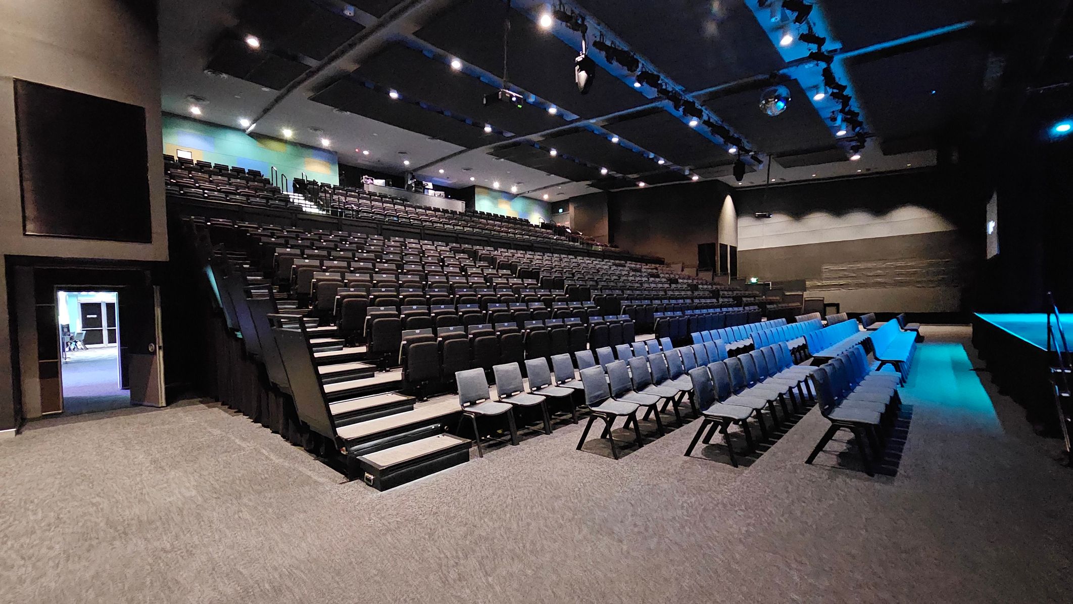 A large auditorium with tiered seating on the left, featuring rows of black chairs. On the right, there are additional rows of chairs on a flat surface. The ceiling is equipped with spotlights and a disco ball, casting a blue hue over the room. The floor is carpeted in grey. A doorway on the far left leads to another room, and the walls are painted in dark tones with some light accents. A large auditorium with tiered seating on the left, featuring rows of black chairs. On the right, there are additional rows of chairs on a flat surface. The ceiling is equipped with spotlights and a disco ball, casting a blue hue over the room. The floor is carpeted in grey. A doorway on the far left leads to another room, and the walls are painted in dark tones with some light accents.