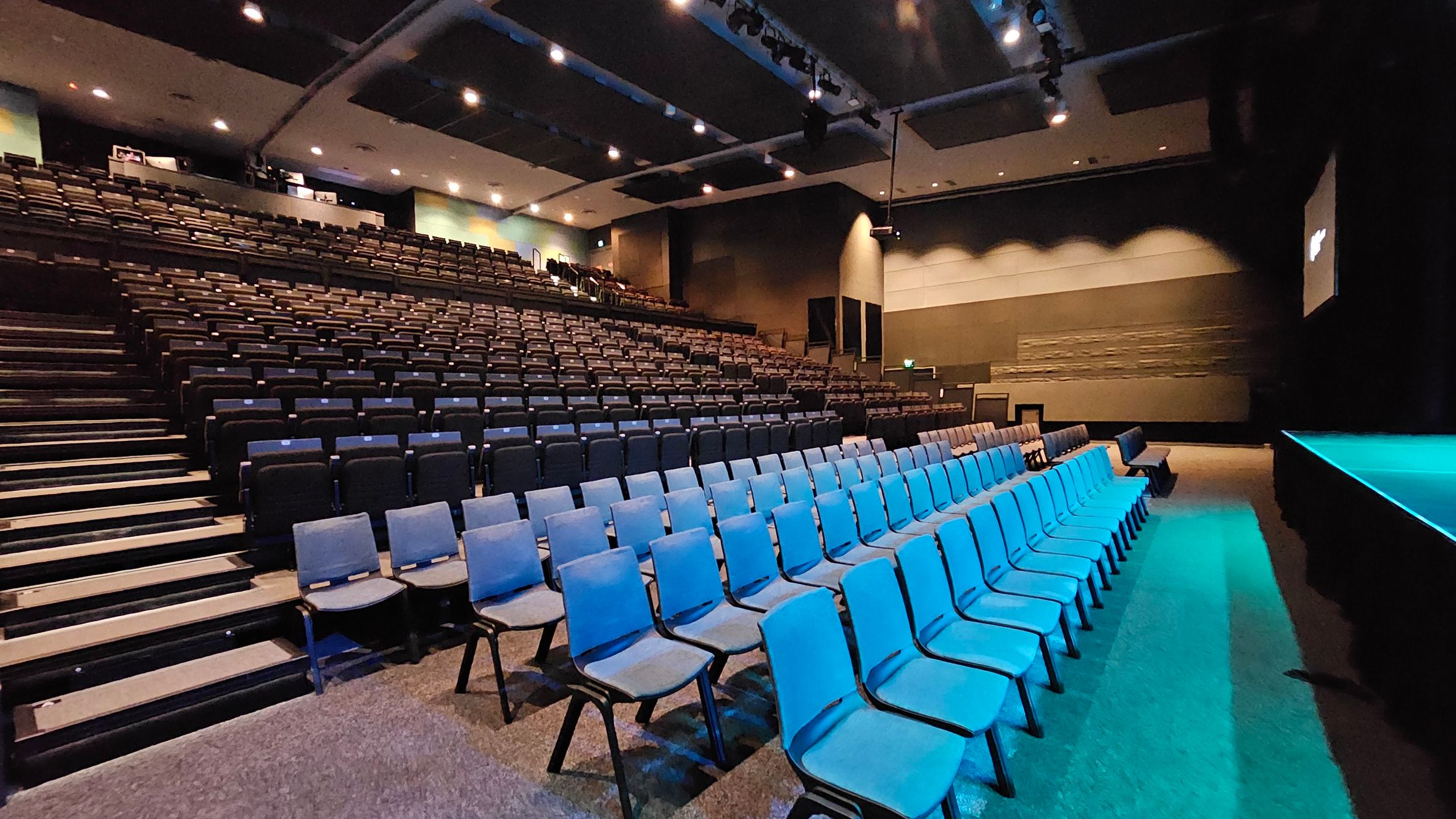 A large auditorium with tiered seating. On the left, rows of dark-coloured seats ascend towards the back. On the right, there are several rows of blue chairs on a flat surface. The stage is visible on the far right, illuminated with blue lighting. The ceiling is equipped with spotlights and acoustic panels. The walls are a mix of dark and light tones, with some textured sections. The floor is carpeted in a neutral colour. A large auditorium with tiered seating. On the left, rows of dark-coloured seats ascend towards the back. On the right, there are several rows of blue chairs on a flat surface. The stage is visible on the far right, illuminated with blue lighting. The ceiling is equipped with spotlights and acoustic panels. The walls are a mix of dark and light tones, with some textured sections. The floor is carpeted in a neutral colour.