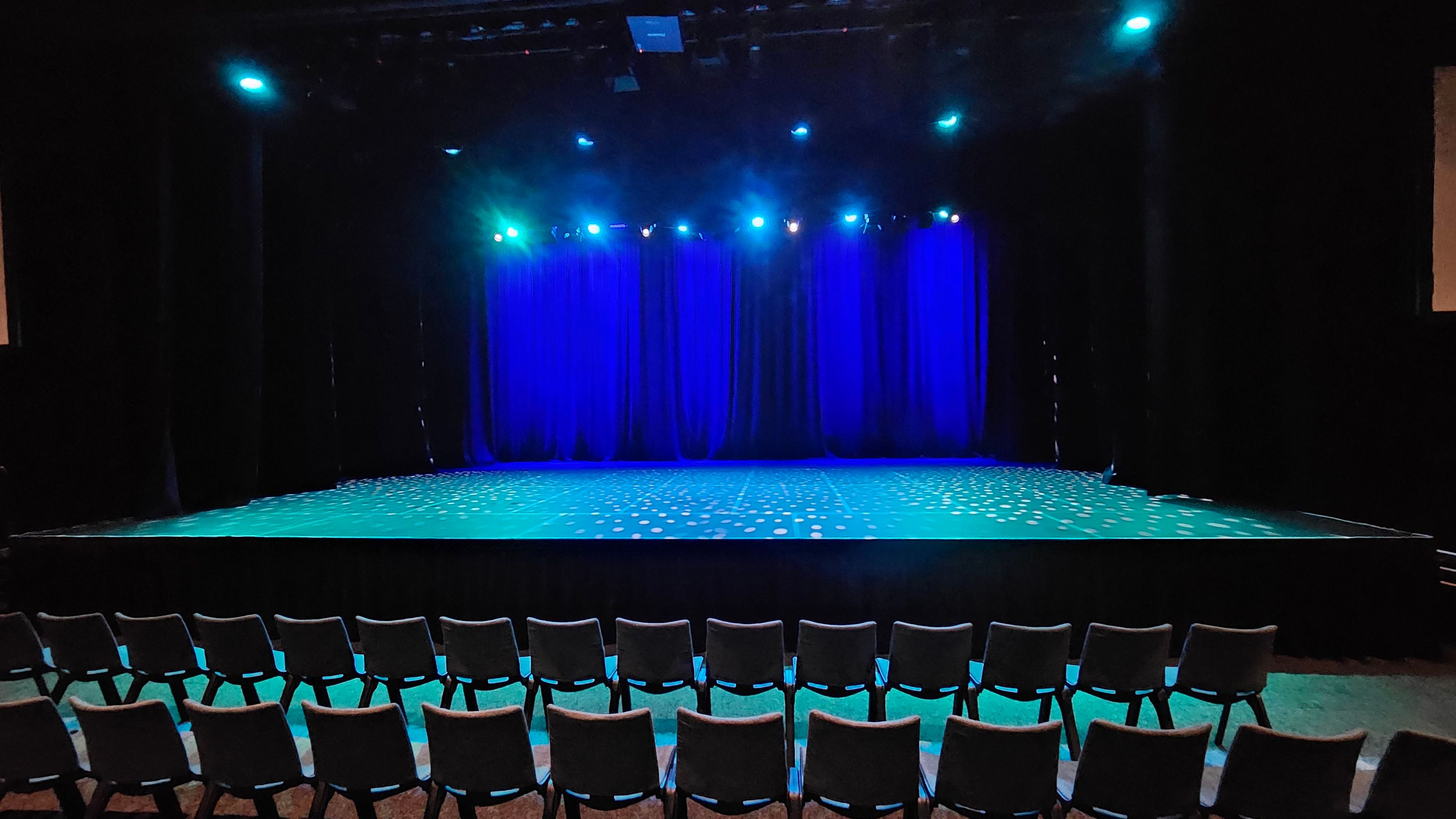 A theatre stage with a row of empty chairs in the foreground. The stage is illuminated with blue lighting, and the floor has a pattern of small white dots. Blue curtains hang in the background, and several spotlights are directed towards the stage. The ceiling is dark, with visible lighting fixtures. The overall atmosphere is dimly lit, with a focus on the stage area. A theatre stage with a row of empty chairs in the foreground. The stage is illuminated with blue lighting, and the floor has a pattern of small white dots. Blue curtains hang in the background, and several spotlights are directed towards the stage. The ceiling is dark, with visible lighting fixtures. The overall atmosphere is dimly lit, with a focus on the stage area.