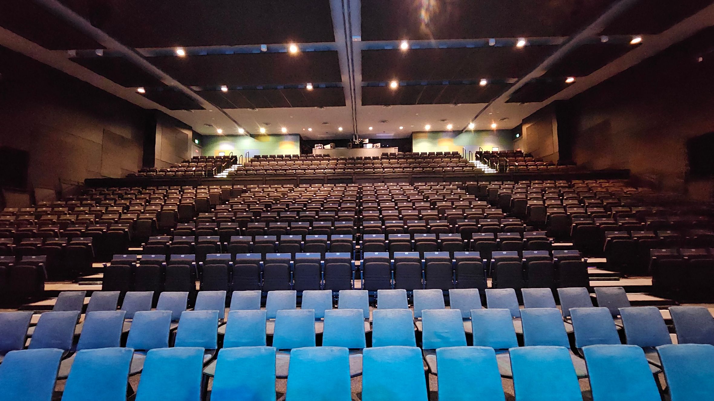 A large auditorium with rows of empty seats, predominantly in dark blue, facing the front. The seating is arranged in a tiered fashion, ascending towards the back. The ceiling is dark with evenly spaced recessed lighting. The walls are dark, and there are exits visible on either side towards the back. The floor is carpeted, and the overall atmosphere is spacious and organised. A large auditorium with rows of empty seats, predominantly in dark blue, facing the front. The seating is arranged in a tiered fashion, ascending towards the back. The ceiling is dark with evenly spaced recessed lighting. The walls are dark, and there are exits visible on either side towards the back. The floor is carpeted, and the overall atmosphere is spacious and organised.