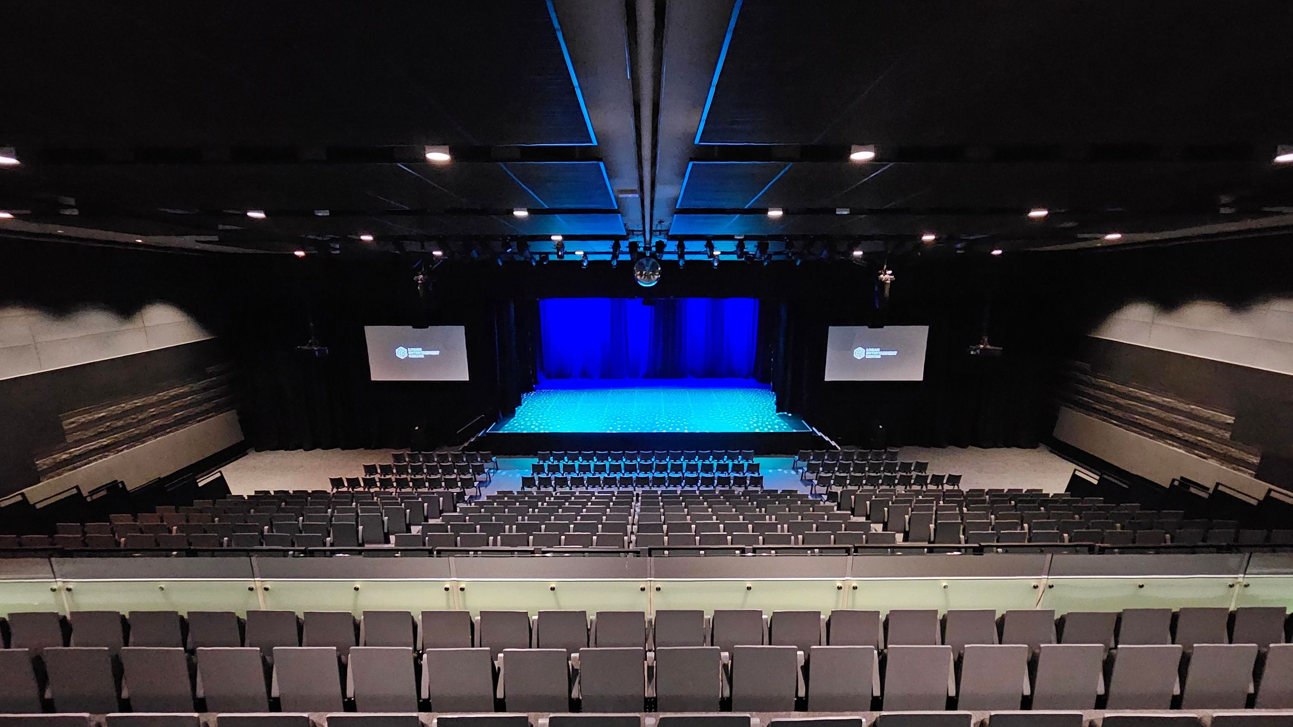 A large auditorium with tiered seating in the foreground, leading down to a stage in the middle. The stage is illuminated with blue lighting, and there are two screens on either side displaying text. The walls are dark with some decorative elements. The ceiling is equipped with multiple lights and a central beam. The floor is carpeted, and the overall atmosphere is set for a performance or presentation. A large auditorium with tiered seating in the foreground, leading down to a stage in the middle. The stage is illuminated with blue lighting, and there are two screens on either side displaying text. The walls are dark with some decorative elements. The ceiling is equipped with multiple lights and a central beam. The floor is carpeted, and the overall atmosphere is set for a performance or presentation.