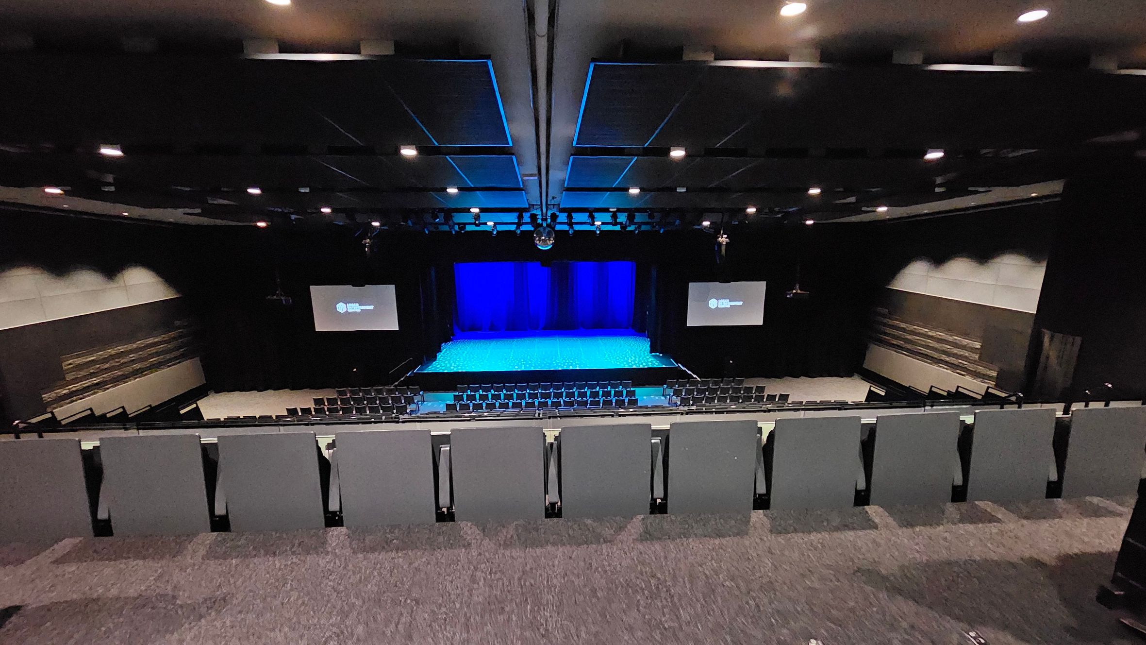 A large auditorium with tiered seating in the foreground, covered in dark fabric. The stage is in the centre, illuminated with blue lighting, and flanked by two large screens displaying text. The walls are lined with acoustic panels, and the ceiling features recessed lighting and sound equipment. The floor is carpeted in a dark colour, complementing the overall dimly lit atmosphere. A large auditorium with tiered seating in the foreground, covered in dark fabric. The stage is in the centre, illuminated with blue lighting, and flanked by two large screens displaying text. The walls are lined with acoustic panels, and the ceiling features recessed lighting and sound equipment. The floor is carpeted in a dark colour, complementing the overall dimly lit atmosphere.
